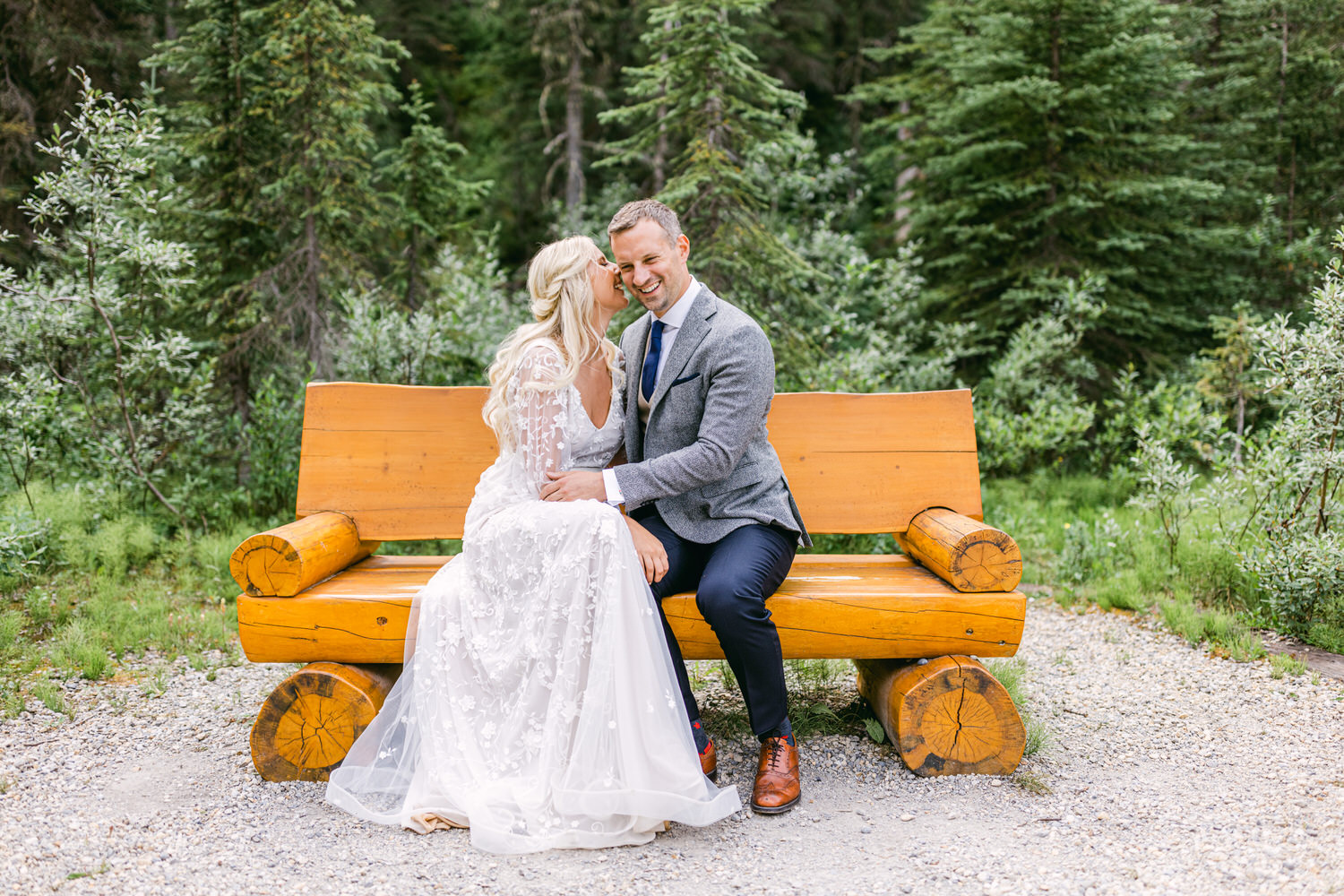 A bride and groom share a playful moment while sitting on a rustic wooden bench in a lush green forest.