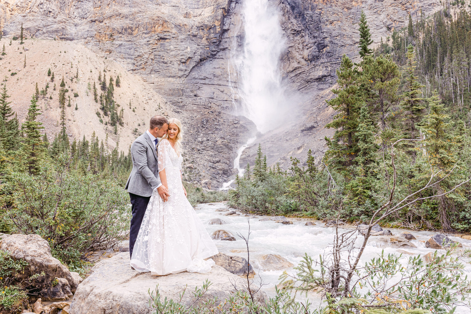 A couple embraces by a river in front of a stunning waterfall, surrounded by lush greenery and rocky mountains.