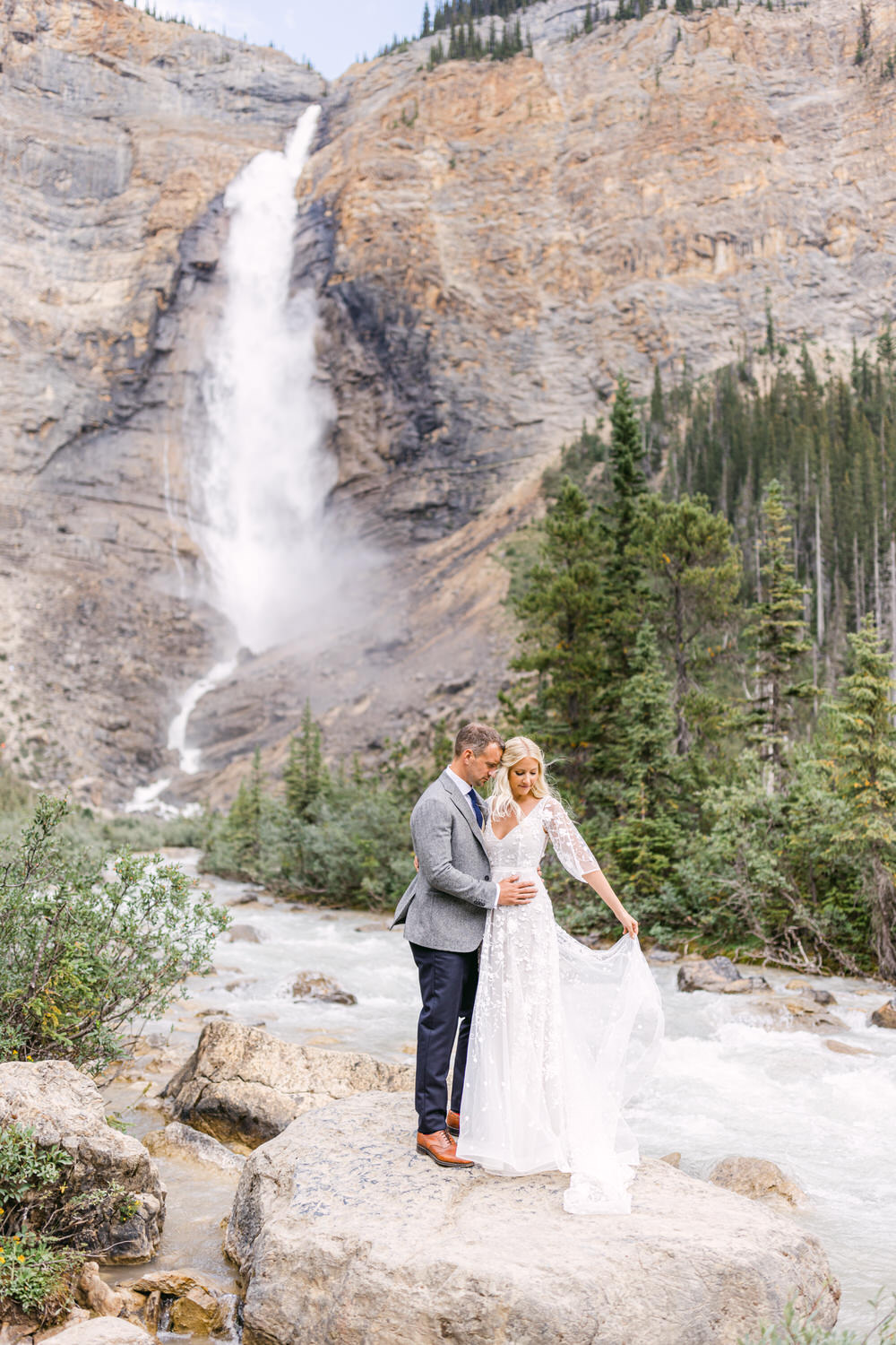 A couple in formal attire stands by a waterfall, surrounded by lush greenery and majestic cliffs, capturing a romantic moment.