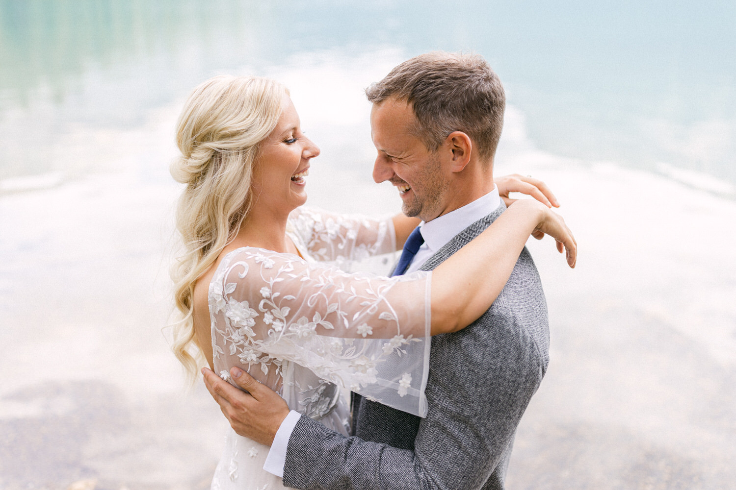 A happy couple sharing a loving moment by a serene water body, with the bride in an elegant dress and the groom in a stylish suit, both smiling at each other.