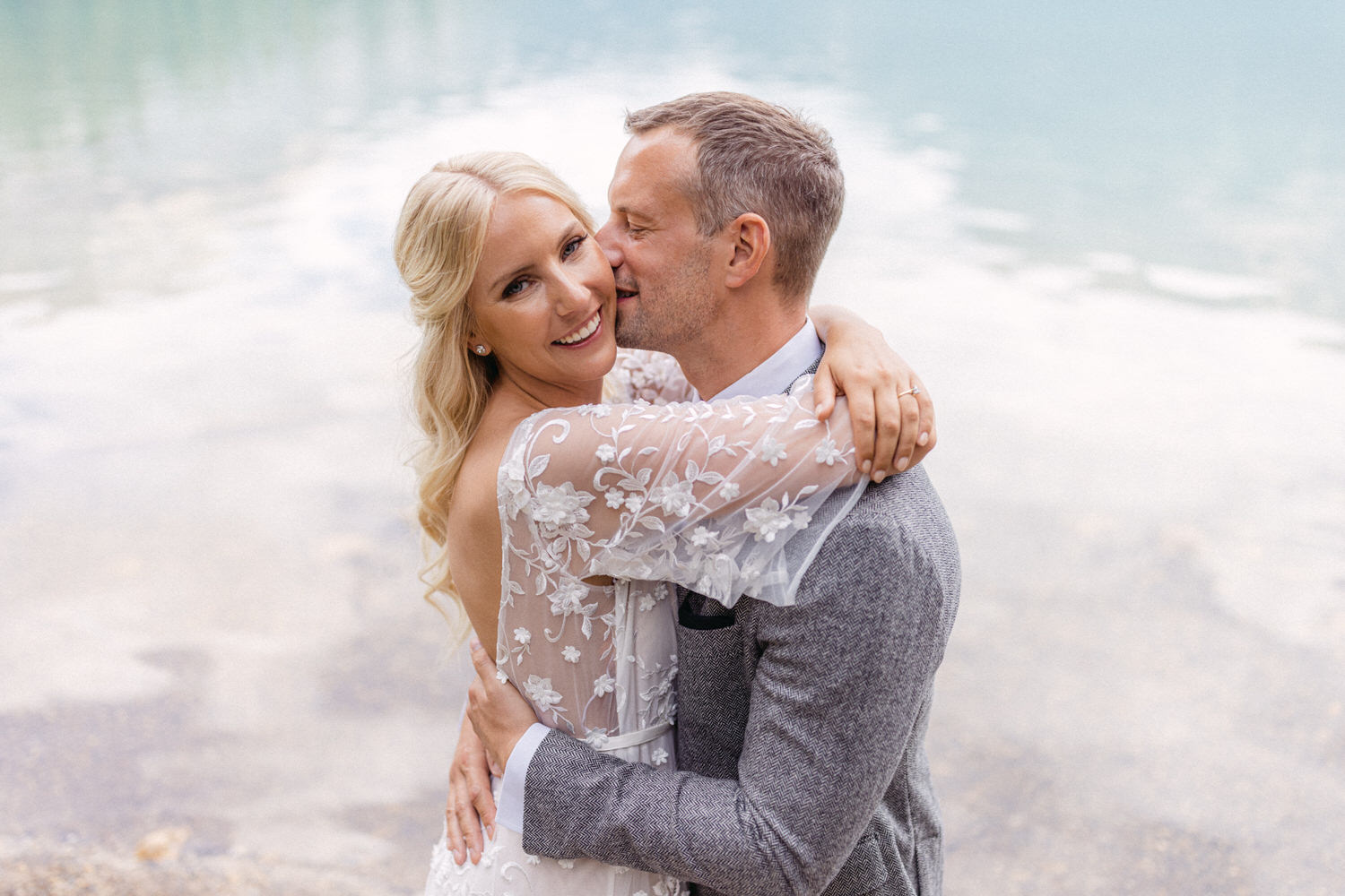 A joyful couple sharing an intimate moment by a serene lake, with the bride in a delicate floral dress and the groom in a smart grey suit.