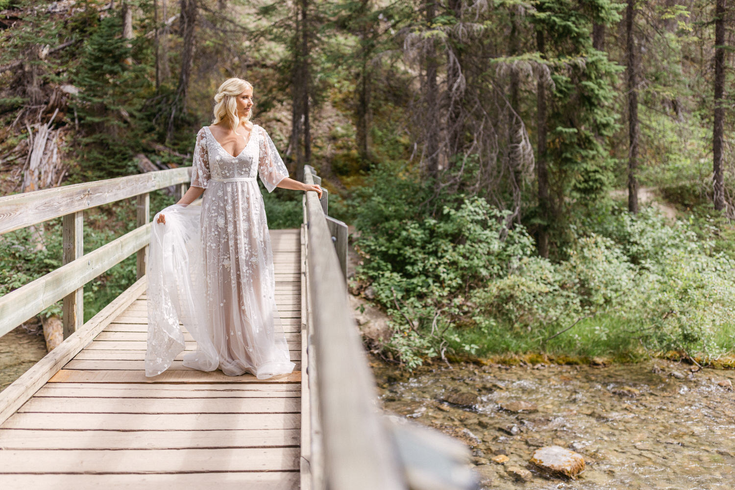 A woman in a flowing, embroidered gown stands gracefully on a wooden bridge surrounded by lush greenery and a serene stream.