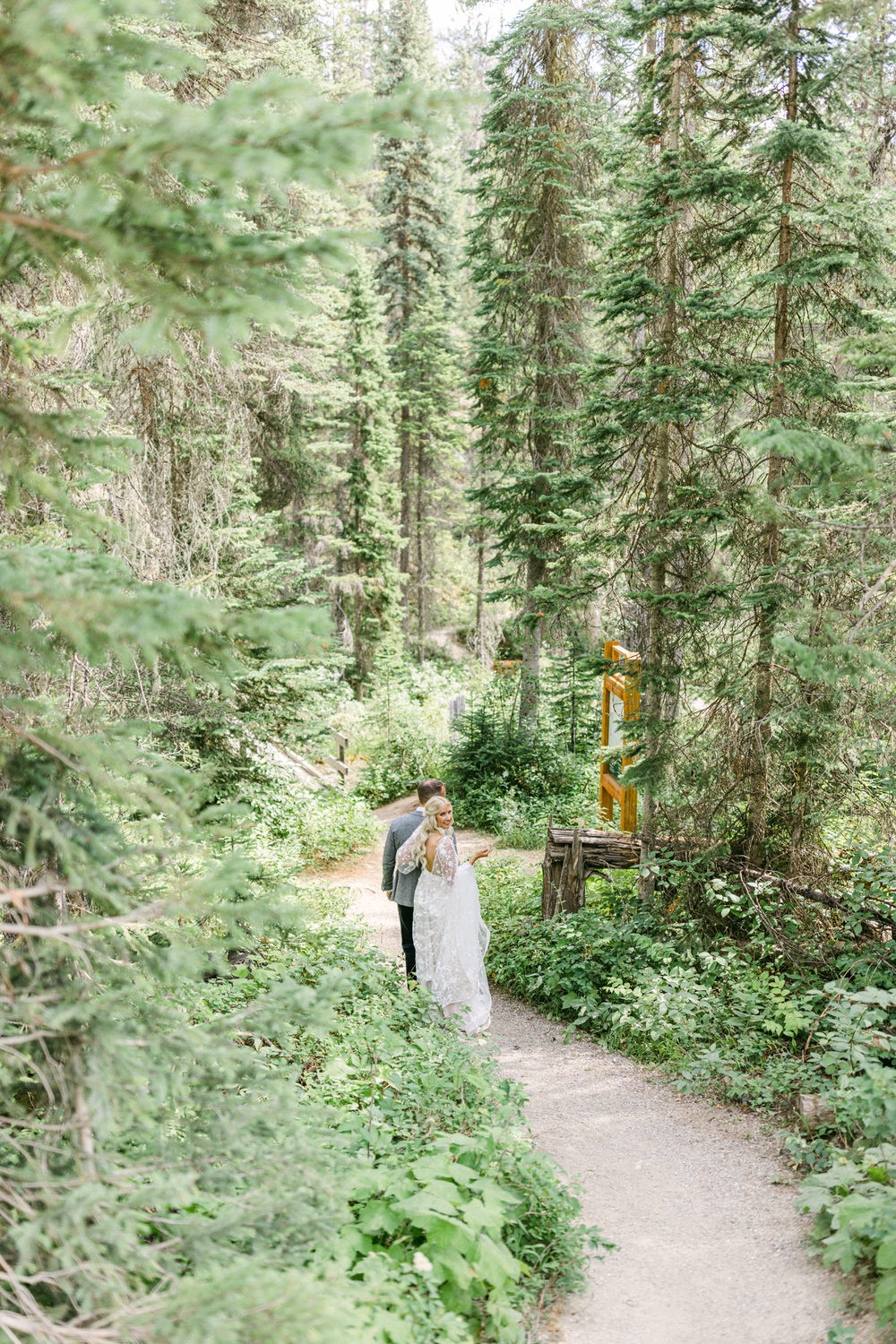 A bride in a lace gown and a groom in a grey suit share a tender moment while walking on a gravel path surrounded by lush greenery and tall trees.