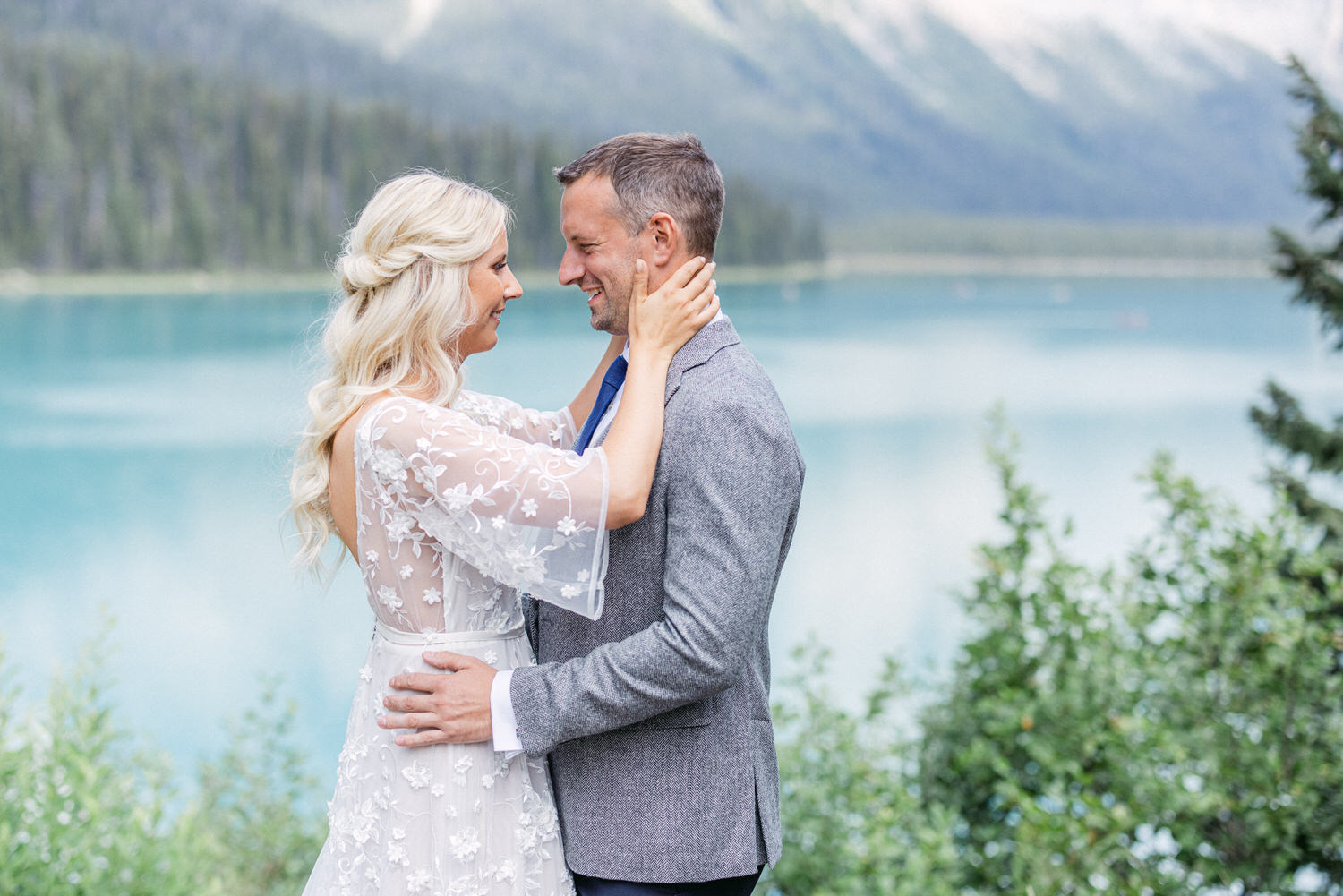 A joyful couple shares an intimate moment by a serene lake, surrounded by lush greenery and majestic mountains. The woman wears a delicate, floral-embellished dress, while the man is in a smart casual outfit, reflecting love and connection.