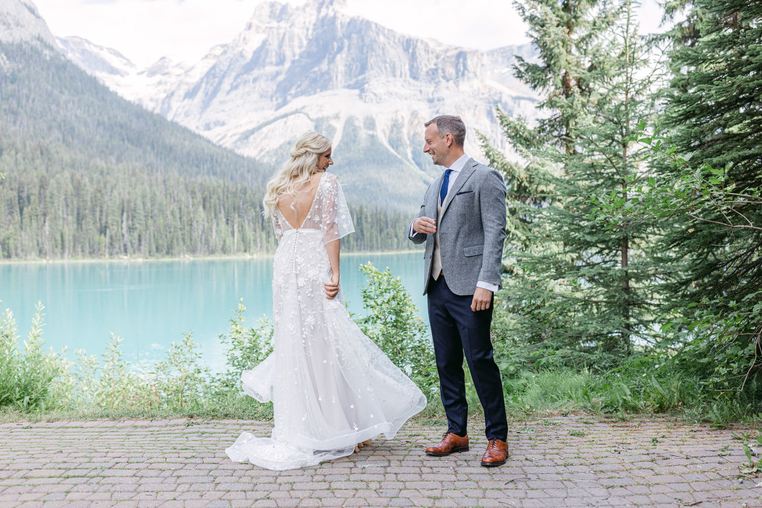 A bride in an elegant, floral-embroidered gown and a groom in a tailored suit stand together near a serene lake, surrounded by mountains and lush greenery.