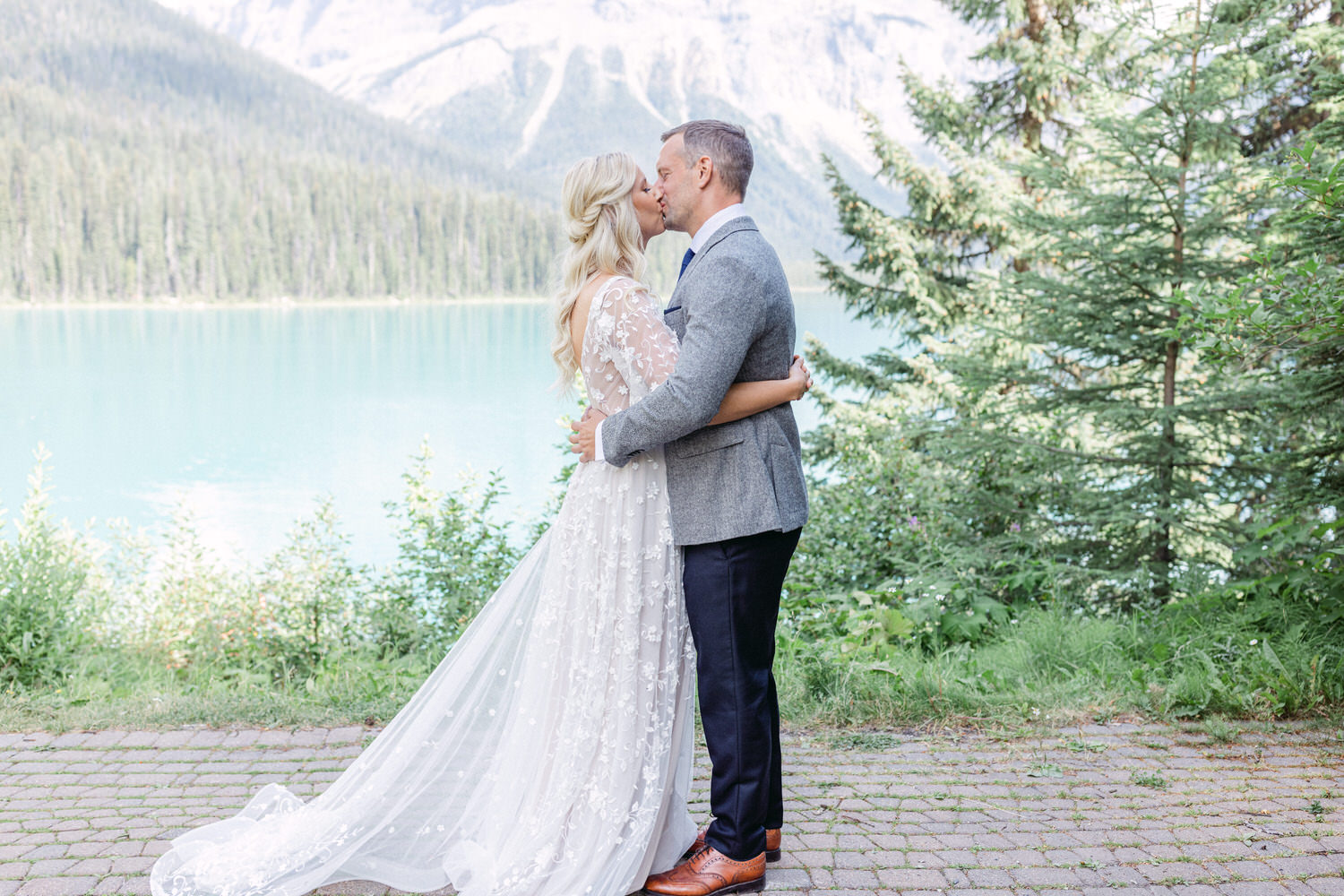 A loving couple sharing a kiss by a serene lake surrounded by lush greenery and mountains.