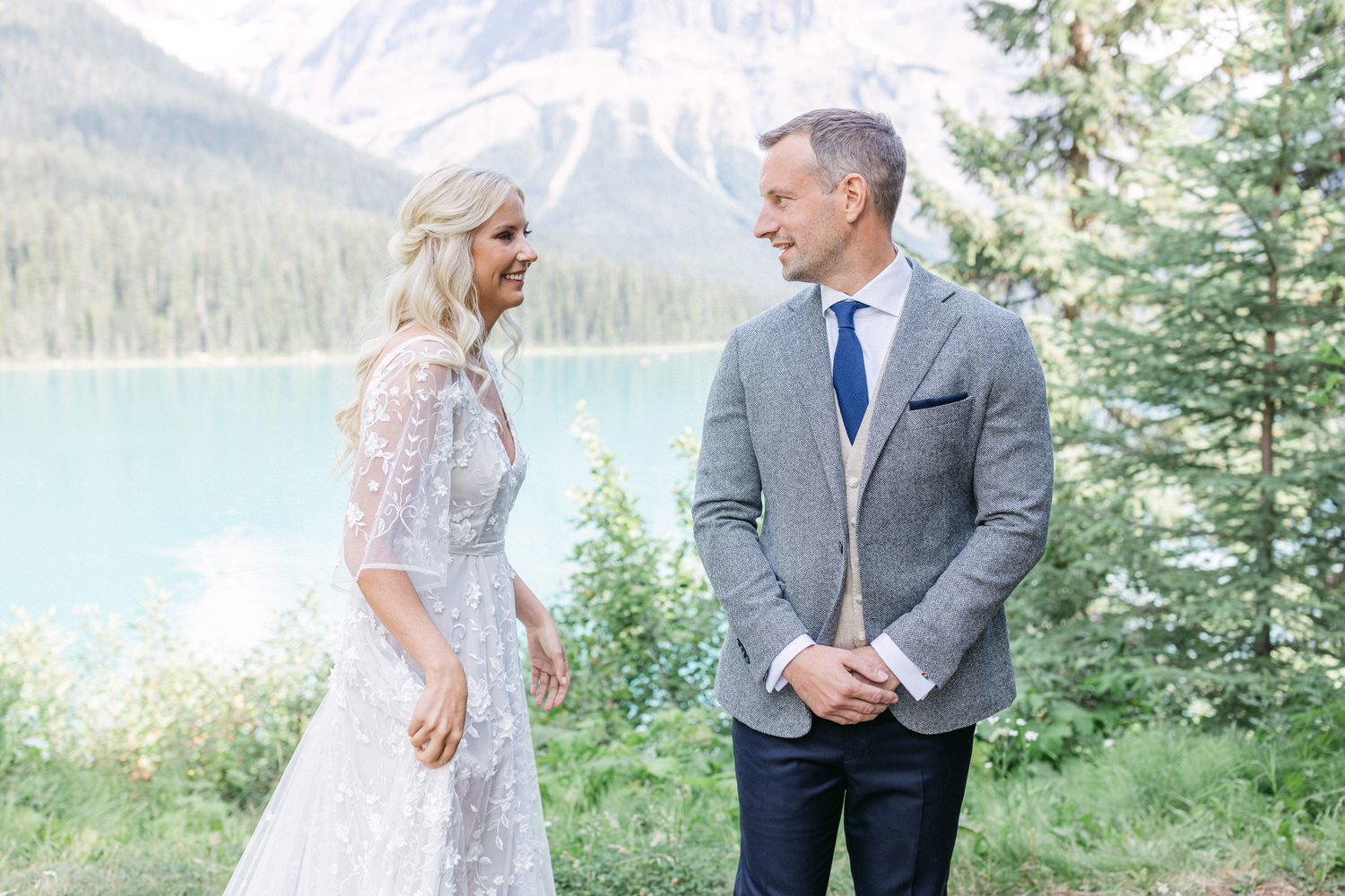 A bride and groom share a joyful smile by a pristine lake surrounded by mountains and greenery.