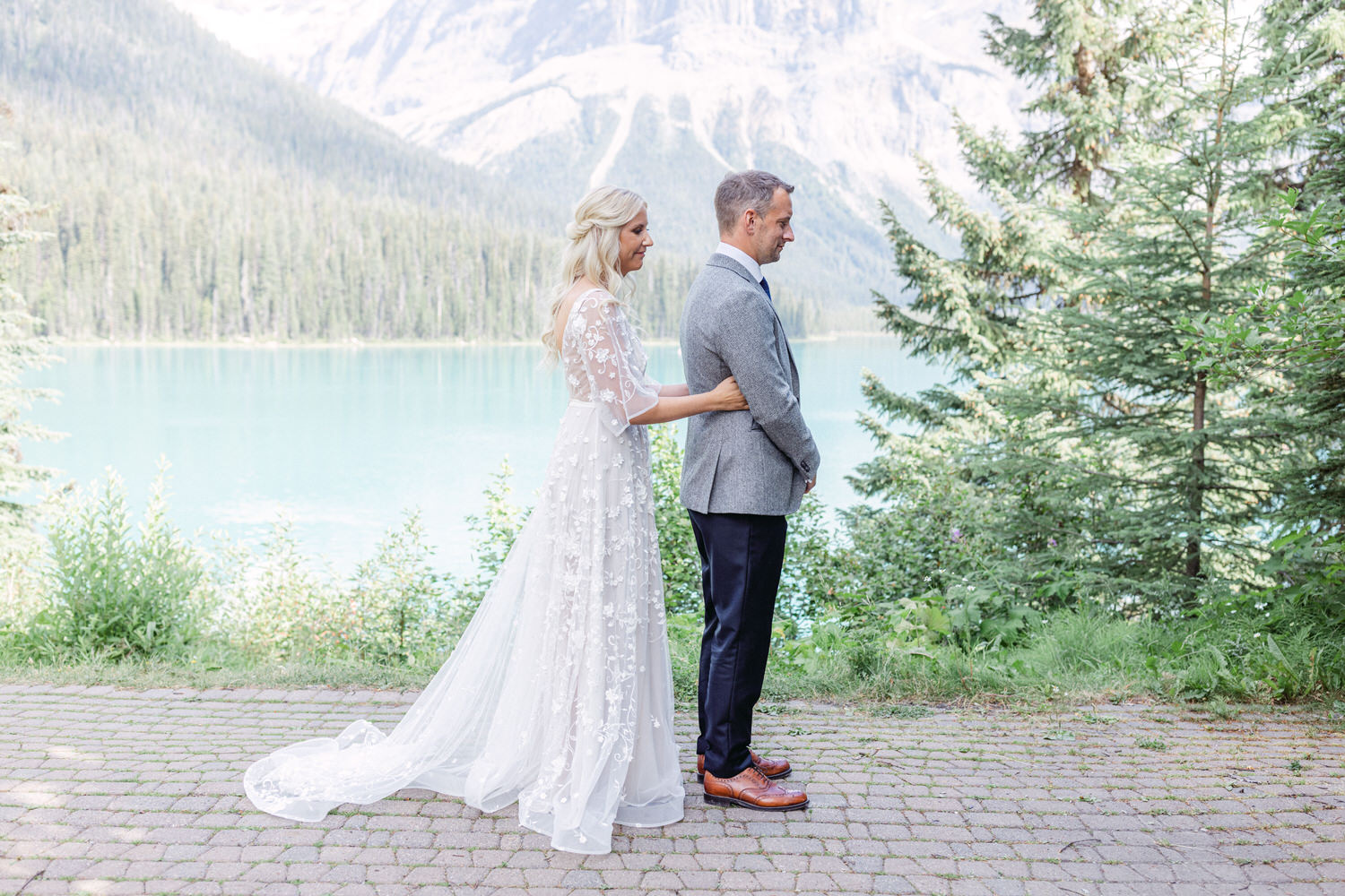 A bride in a floral lace dress embraces her groom, who stands with his back to her, near a serene turquoise lake surrounded by evergreen trees and majestic mountains.