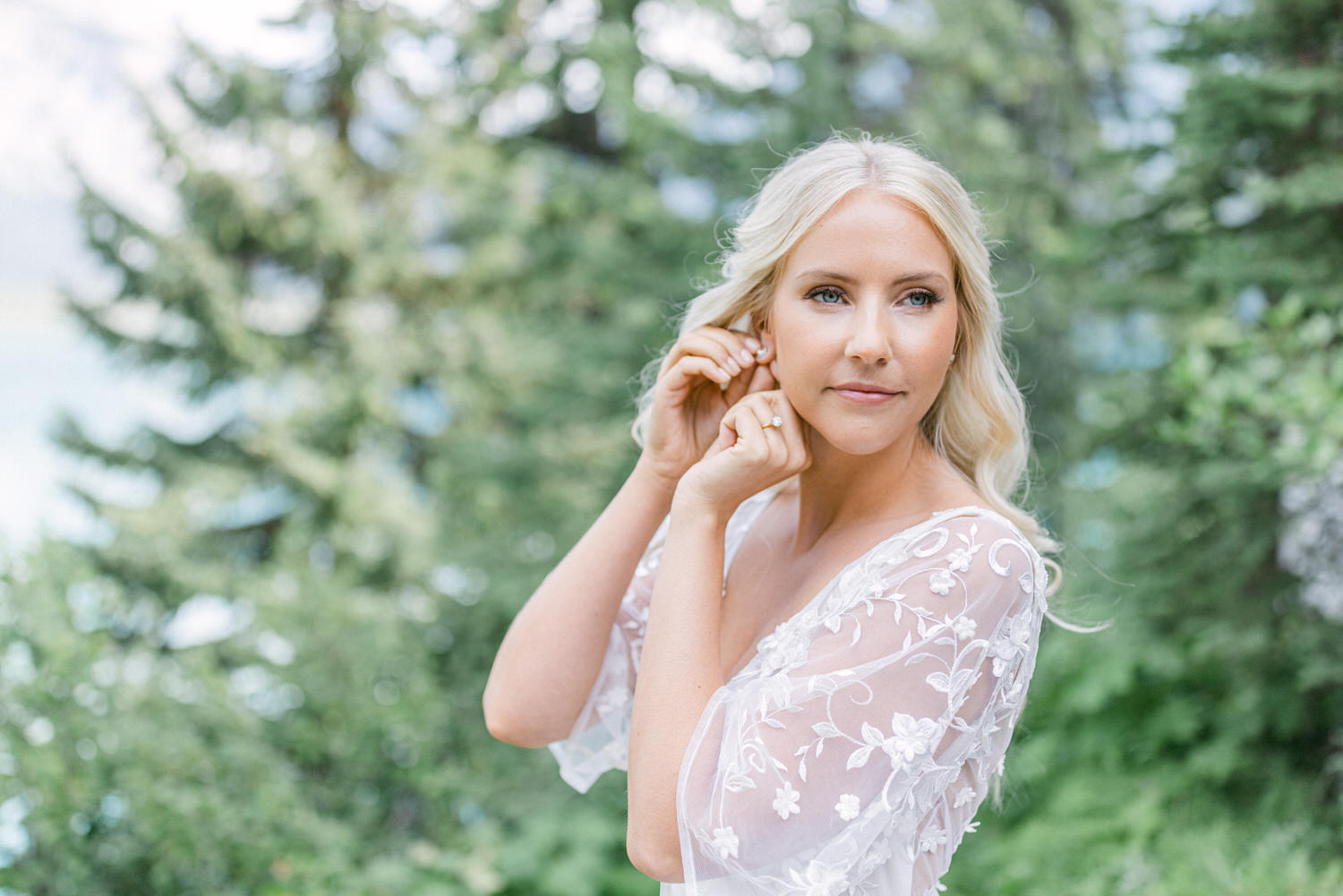 A young woman with long blonde hair poses gently in a lush green setting, wearing a delicate, embroidered white dress and a soft expression, with trees and a serene background.