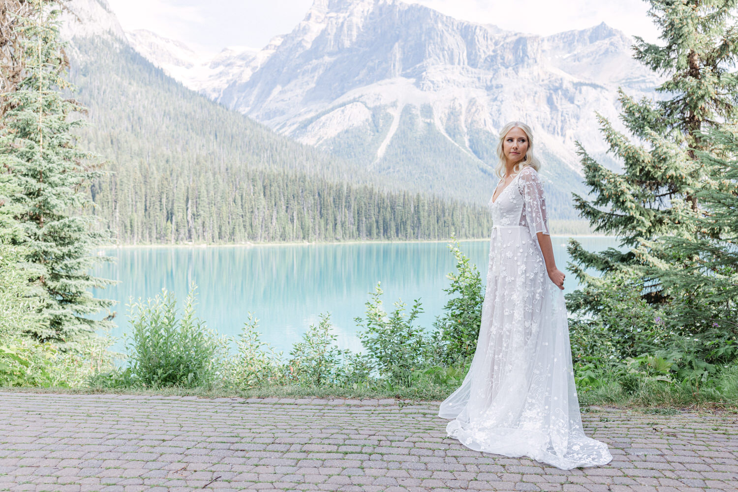 A bride in a delicate white wedding dress stands beside a tranquil mountain lake, surrounded by lush greenery and towering mountains in the background.