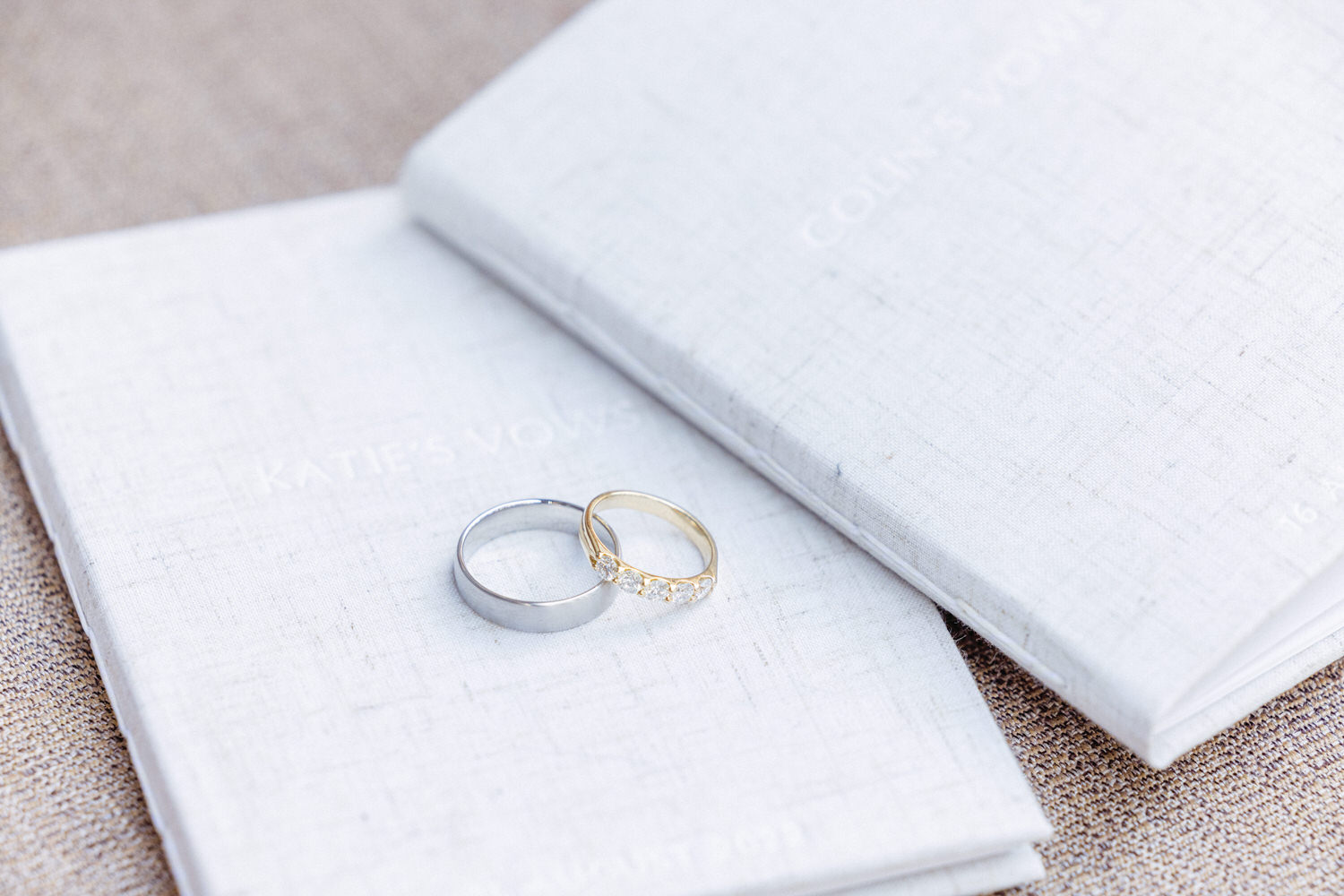 Close-up of a silver wedding band and a gold ring adorned with diamonds, resting atop vow books labeled "Katie's Vows" and "Colin's Vows."