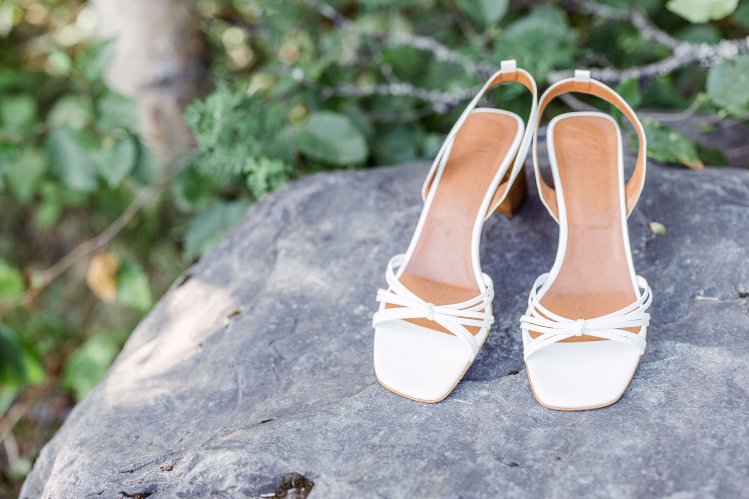 A pair of stylish white open-toe heels resting on a rock, surrounded by green foliage.