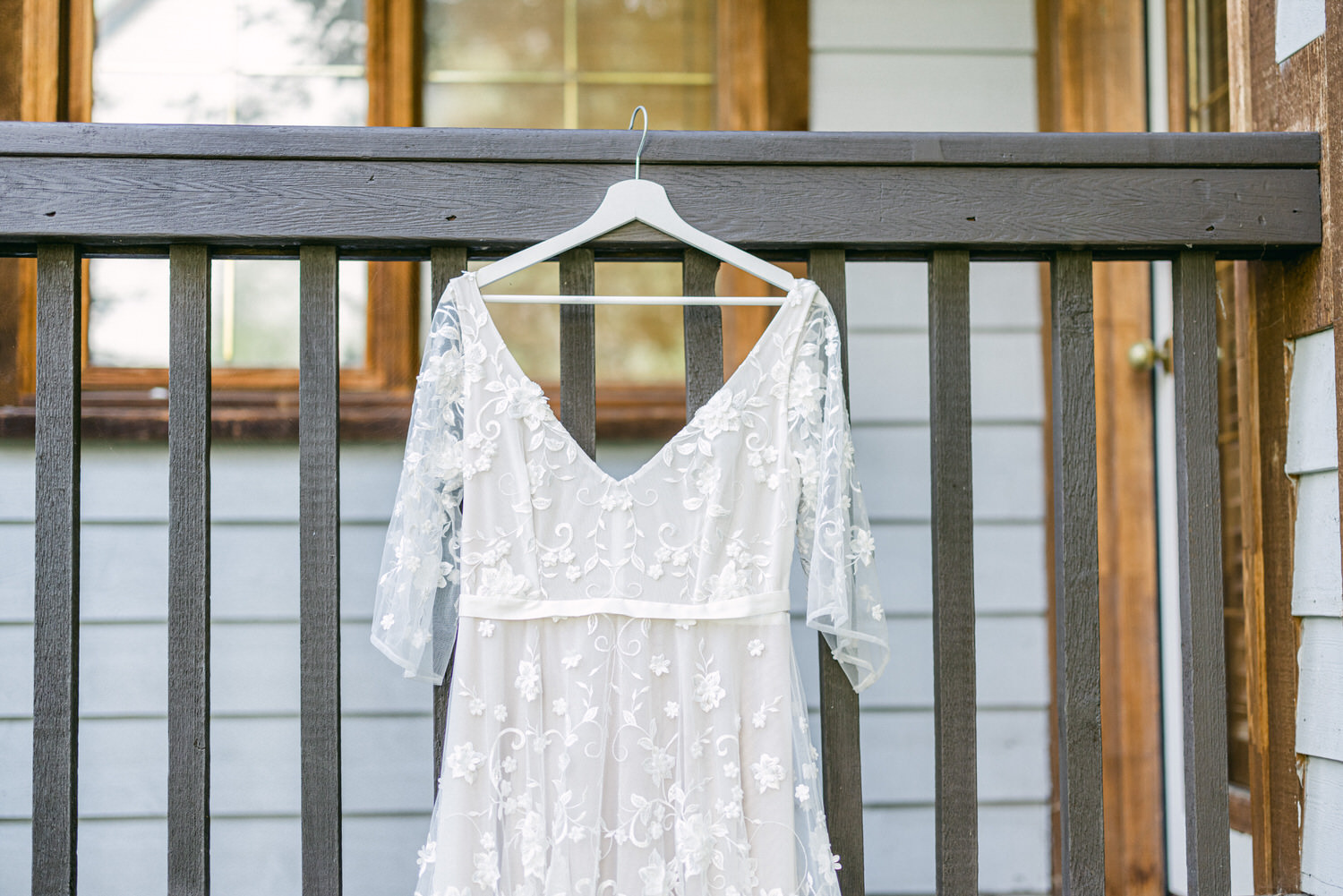 Delicate white wedding dress with floral embroidery hanging on a wooden railing, with a backdrop of a house exterior.