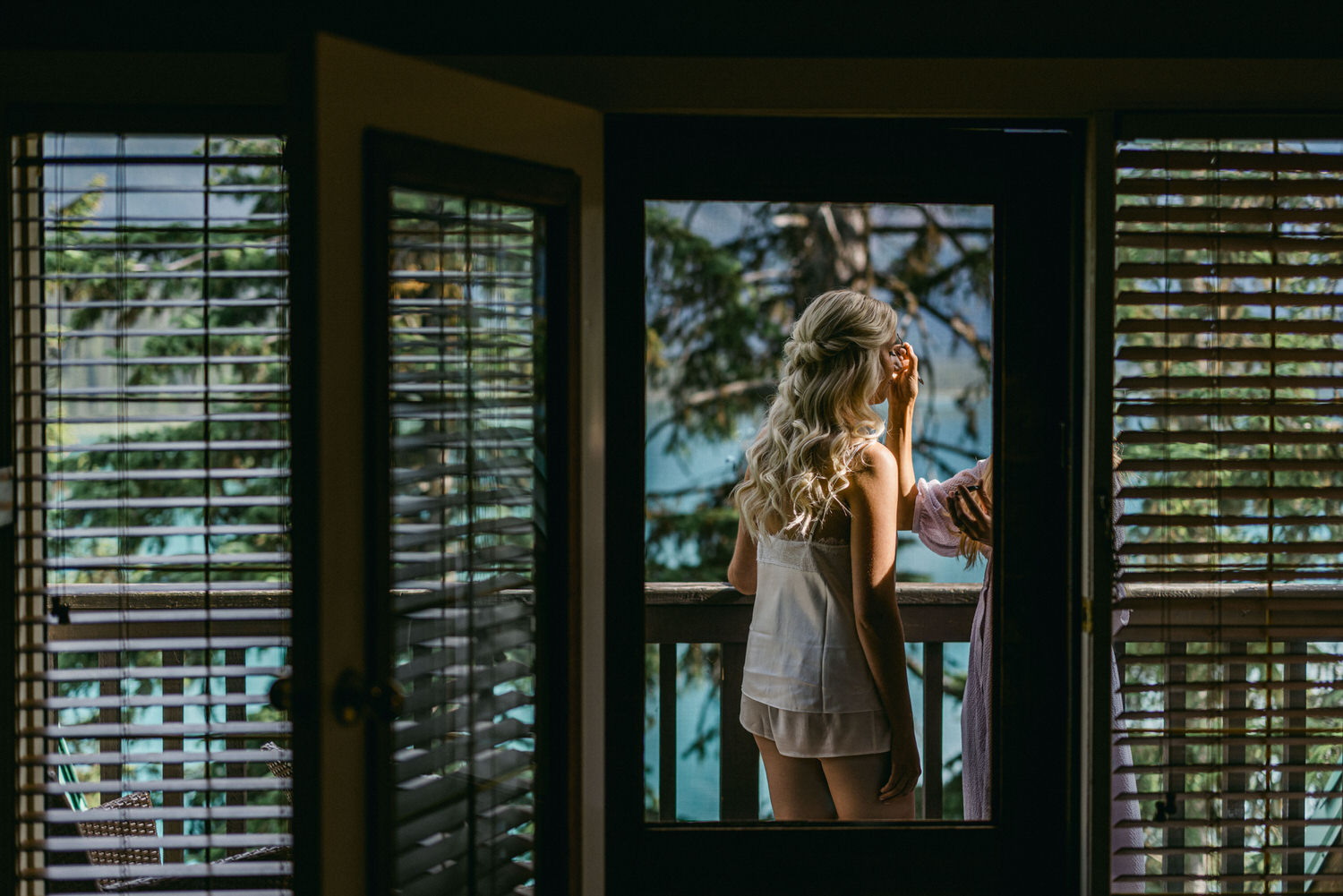 Bridal Preparations in Nature::A woman touches her hair while looking out a patio door, framed by wooden blinds, with a serene forest and lake view.