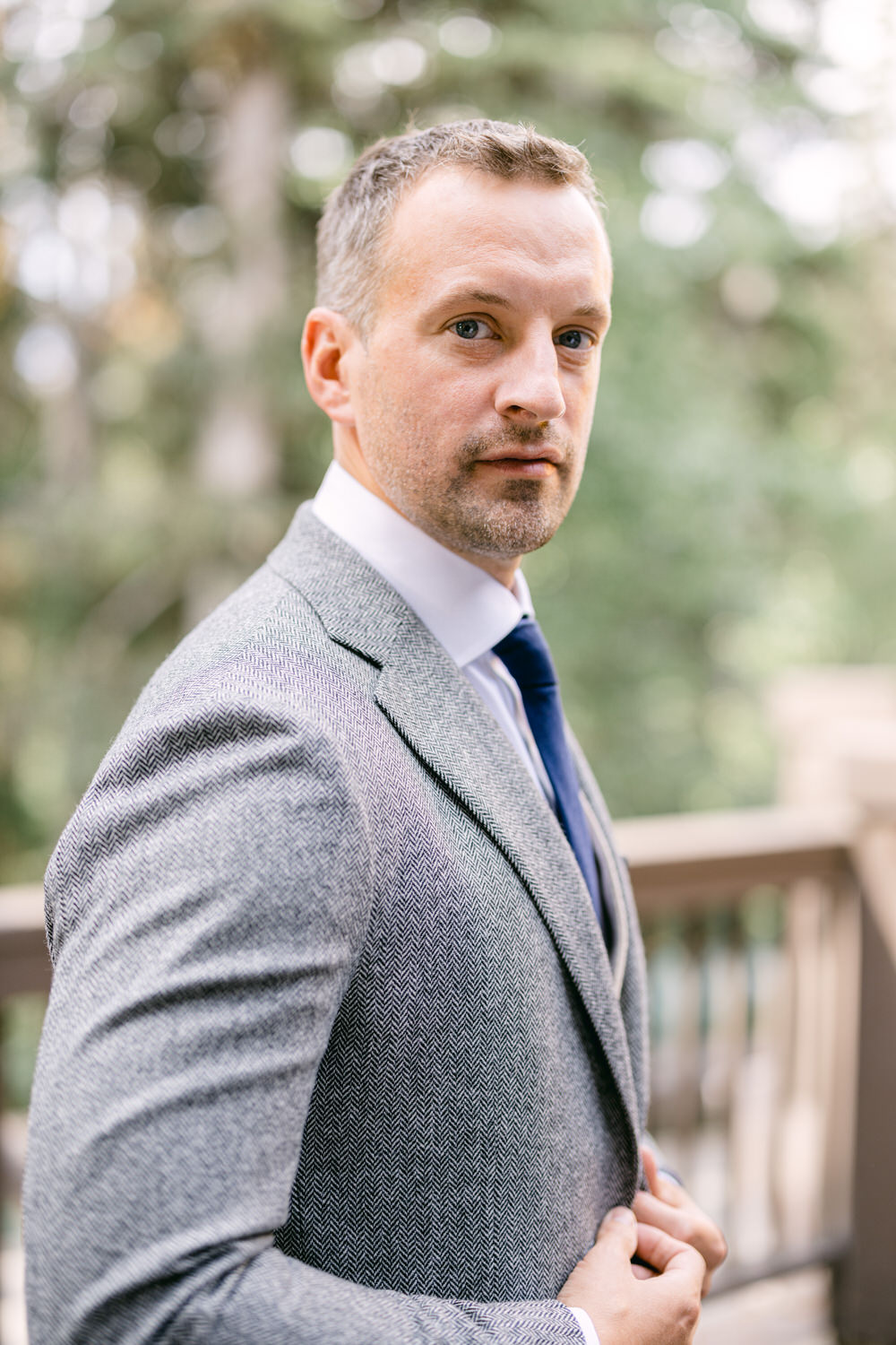 A man in a tailored gray herringbone suit with a navy tie stands confidently outdoors, surrounded by blurred greenery.