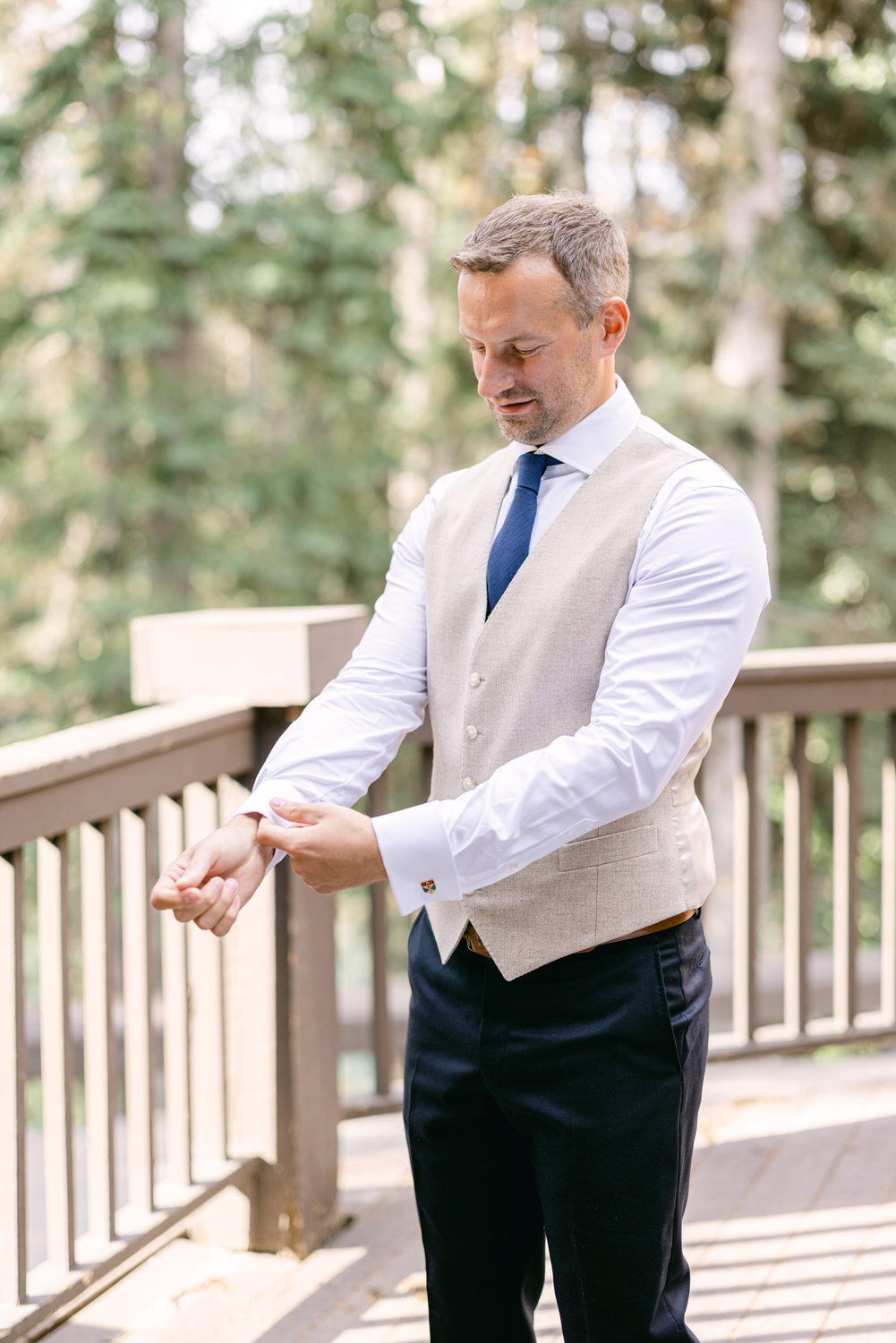 A man in a smart vest adjusts the cuffs of his dress shirt while standing on a wooden deck surrounded by trees.