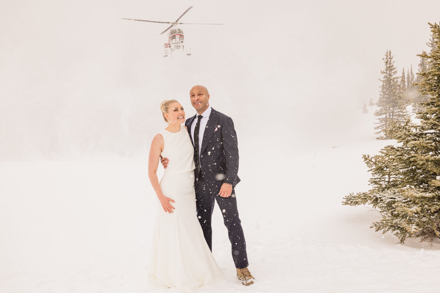 A smiling couple in wedding attire stands together in a snowy landscape, with a helicopter hovering in the background amidst falling snow.