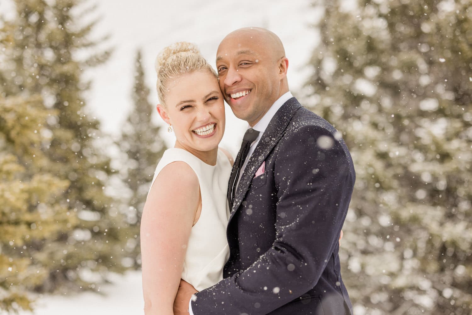 A joyful couple smiling and embracing in a snowy outdoor setting, surrounded by snow-covered trees.