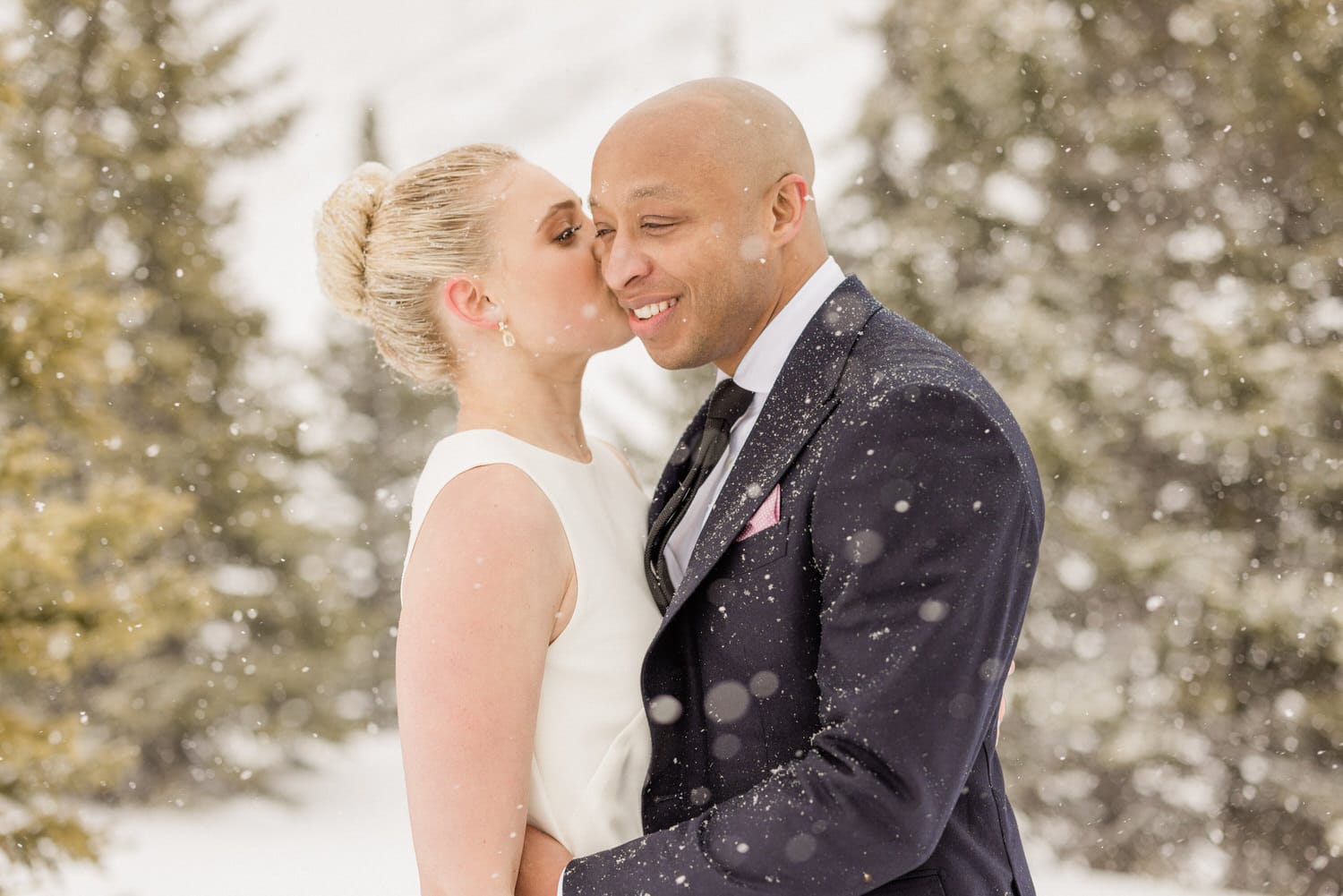 A couple sharing a tender moment in a snowy landscape during their wedding celebration, surrounded by trees and falling snowflakes.