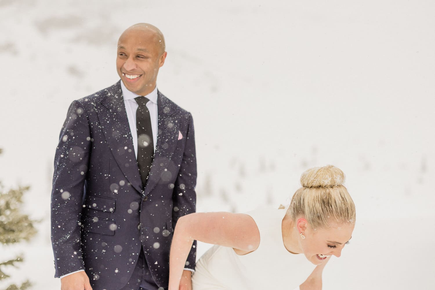 A couple shares a joyful moment in a snowy landscape, with a man in a suit smiling and a woman in a white dress laughing as she bends down.