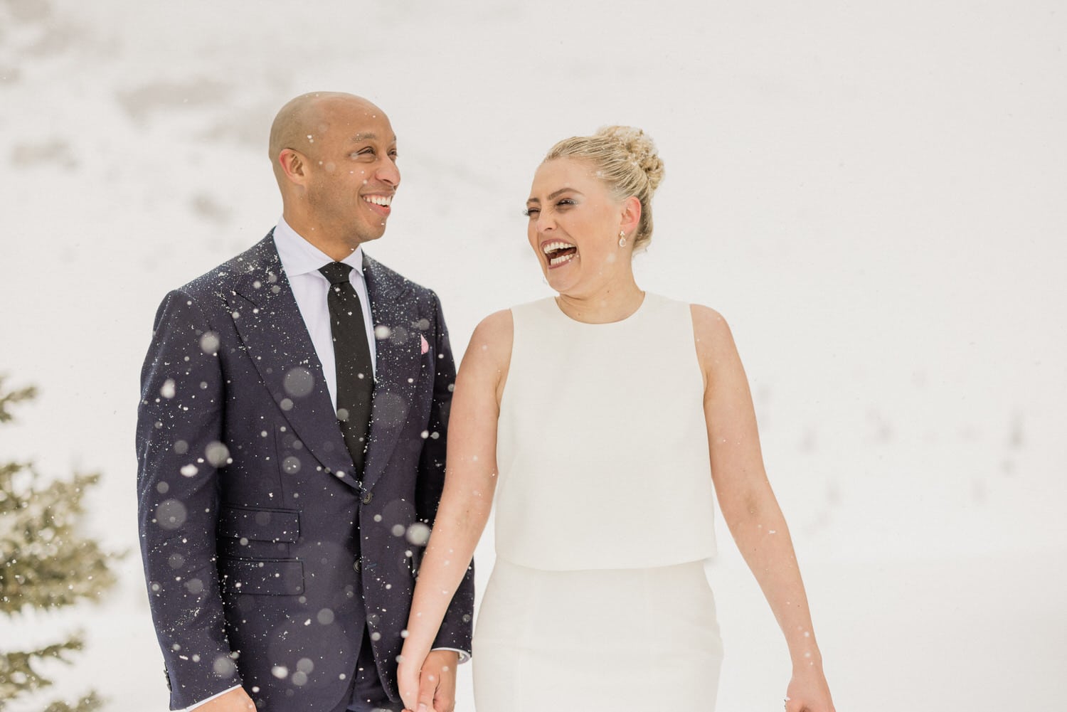 A happy couple holding hands and laughing together in the snow, dressed elegantly for their winter wedding.