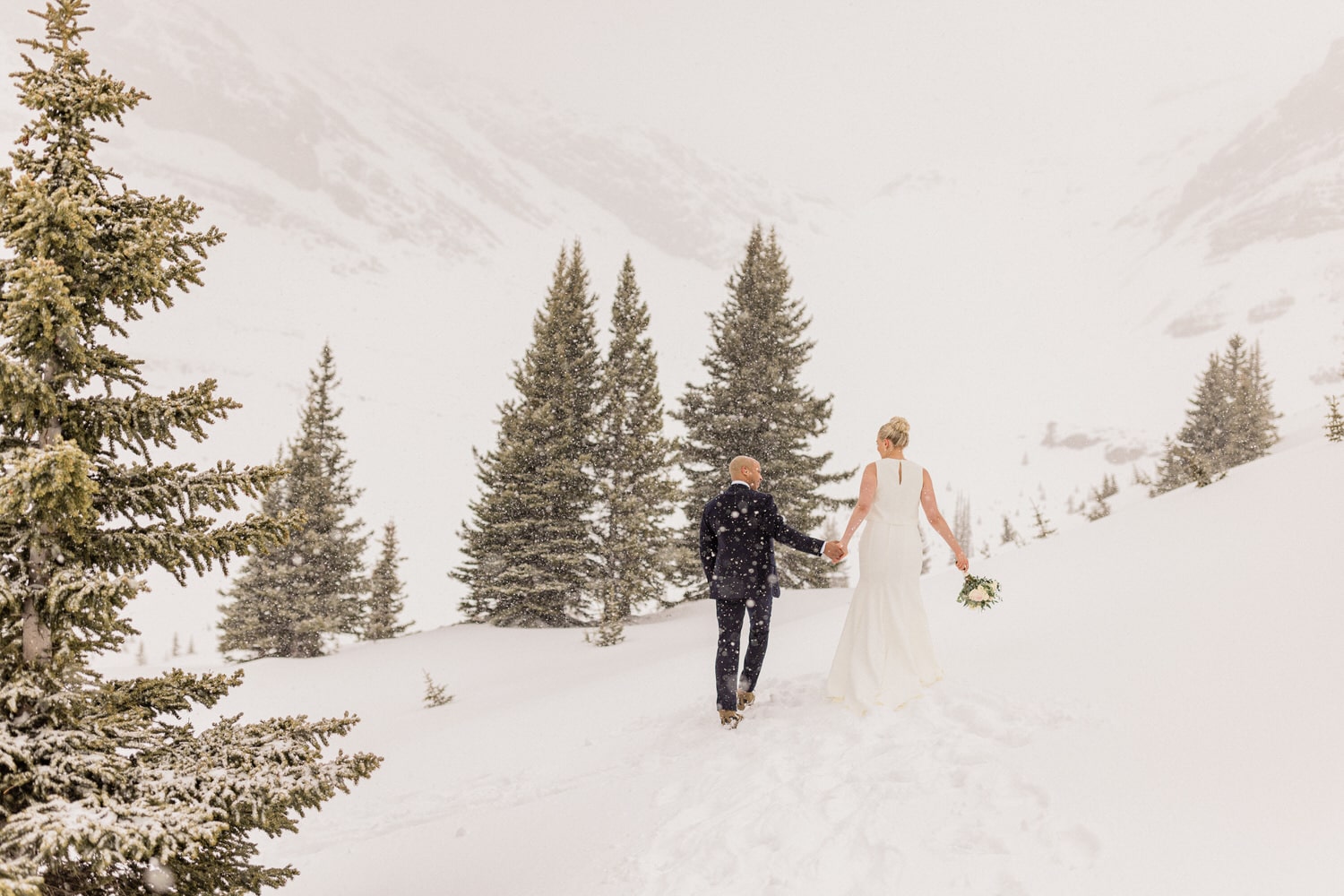 A bride and groom walk hand in hand through a snowy landscape, surrounded by evergreen trees, creating a serene and romantic atmosphere.