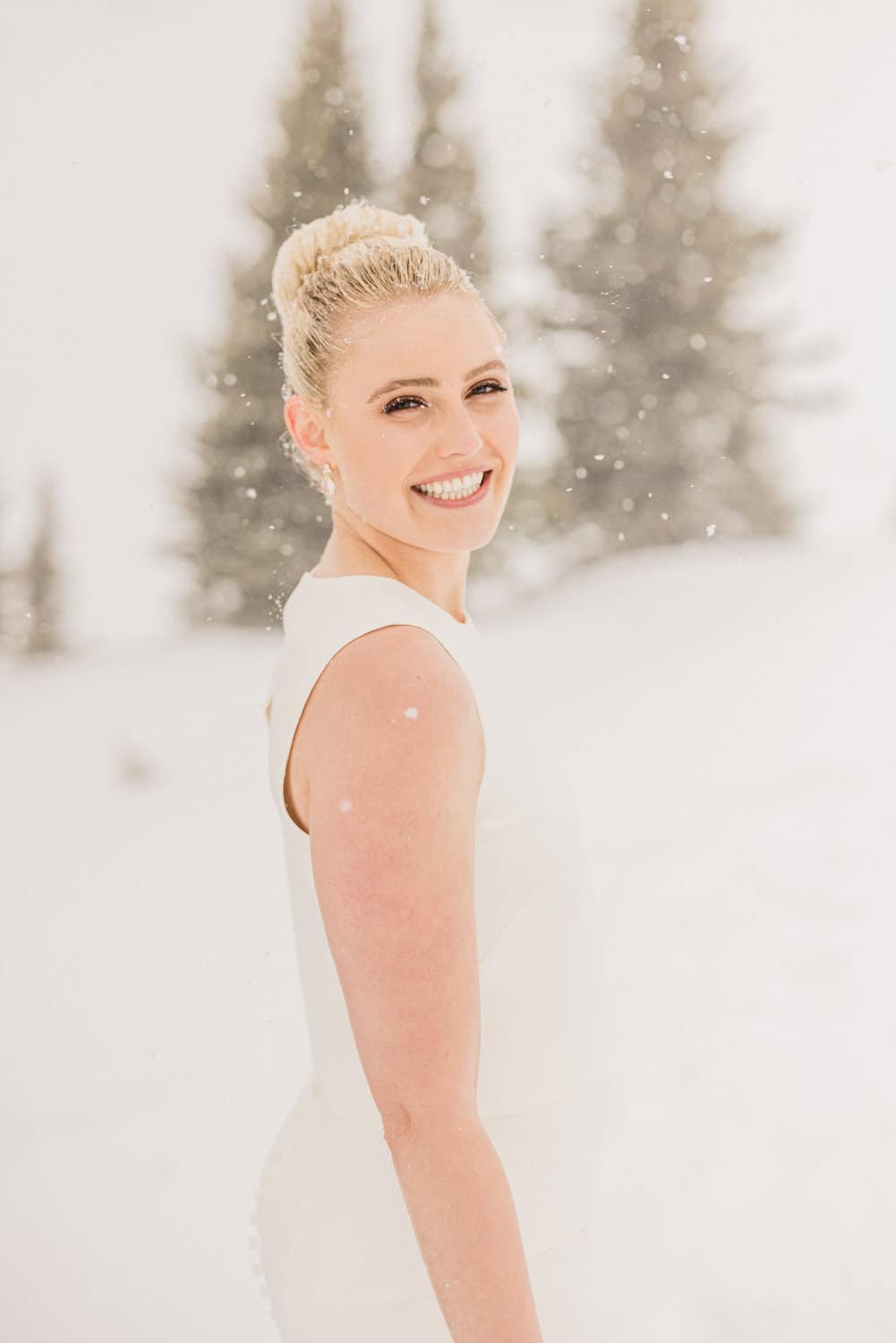 A smiling woman in a white dress stands in a snowy setting, surrounded by evergreens, capturing a joyful moment in winter.