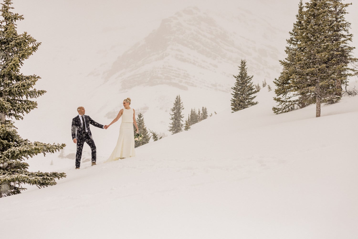 A couple dressed elegantly, holding hands in a snowy landscape, surrounded by evergreen trees and mountains in the background.