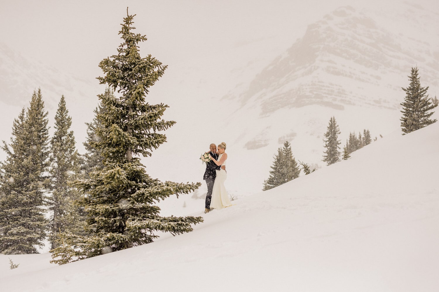 A couple embraces in a snowy landscape, surrounded by evergreen trees and mountains, capturing a romantic moment in winter.