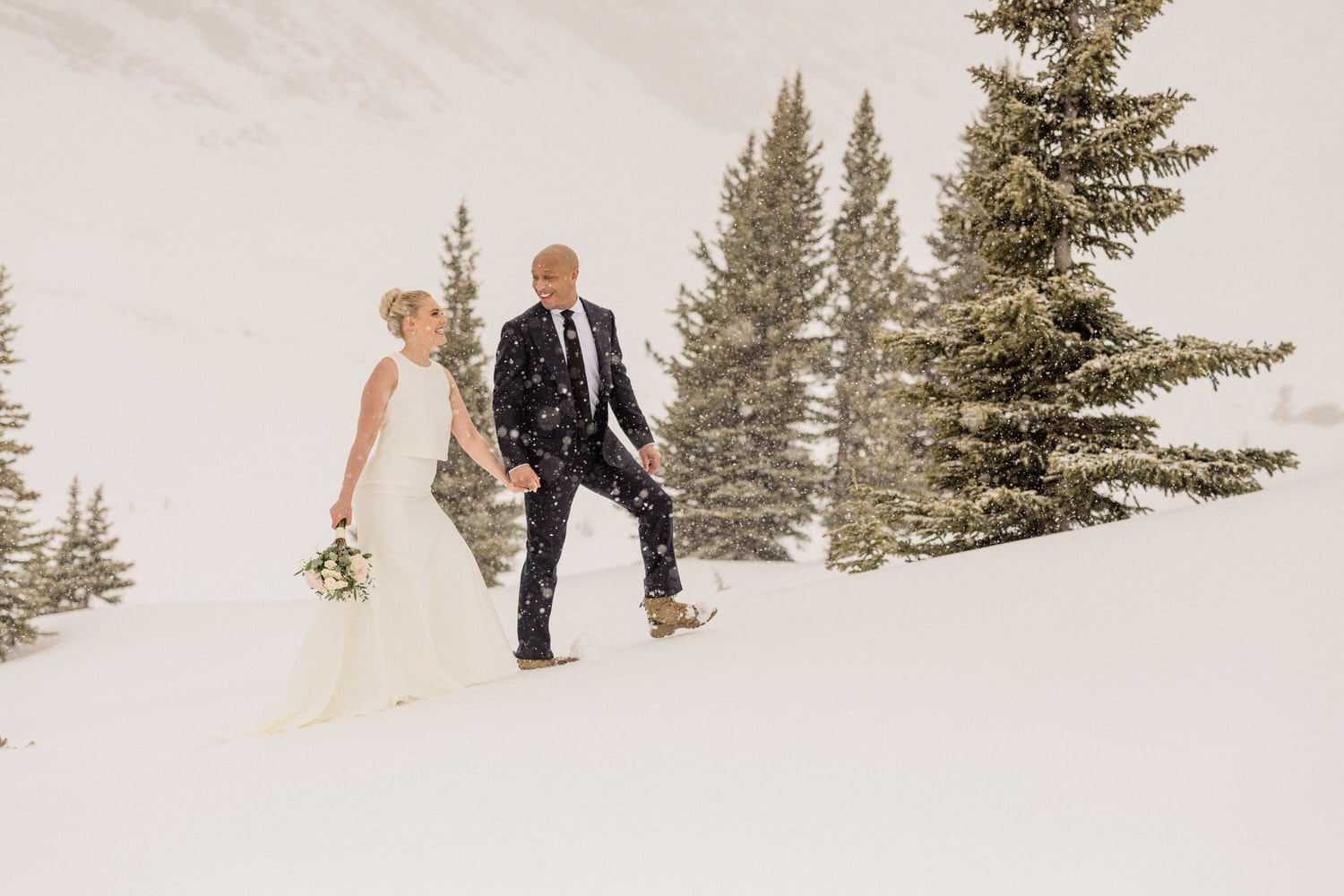 A bride and groom walk hand in hand through a snowy landscape, surrounded by evergreen trees, as snow gently falls around them.