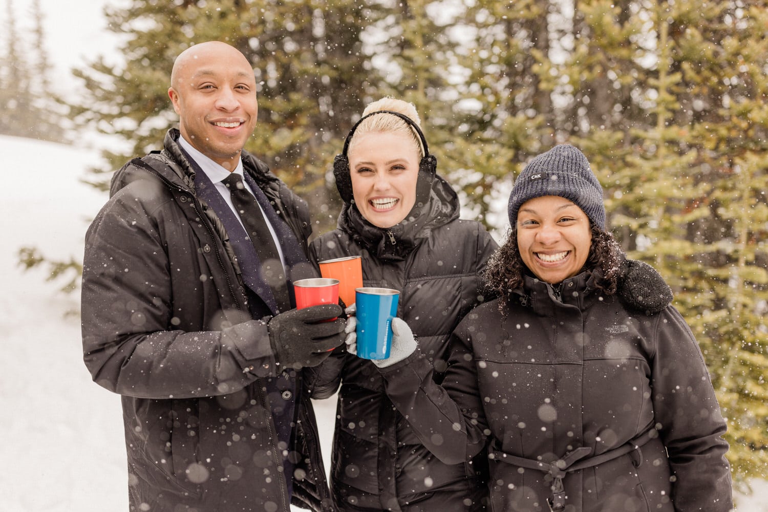 Three friends holding colorful cups while enjoying a snowy outdoor setting, smiling and dressed warmly in winter jackets.