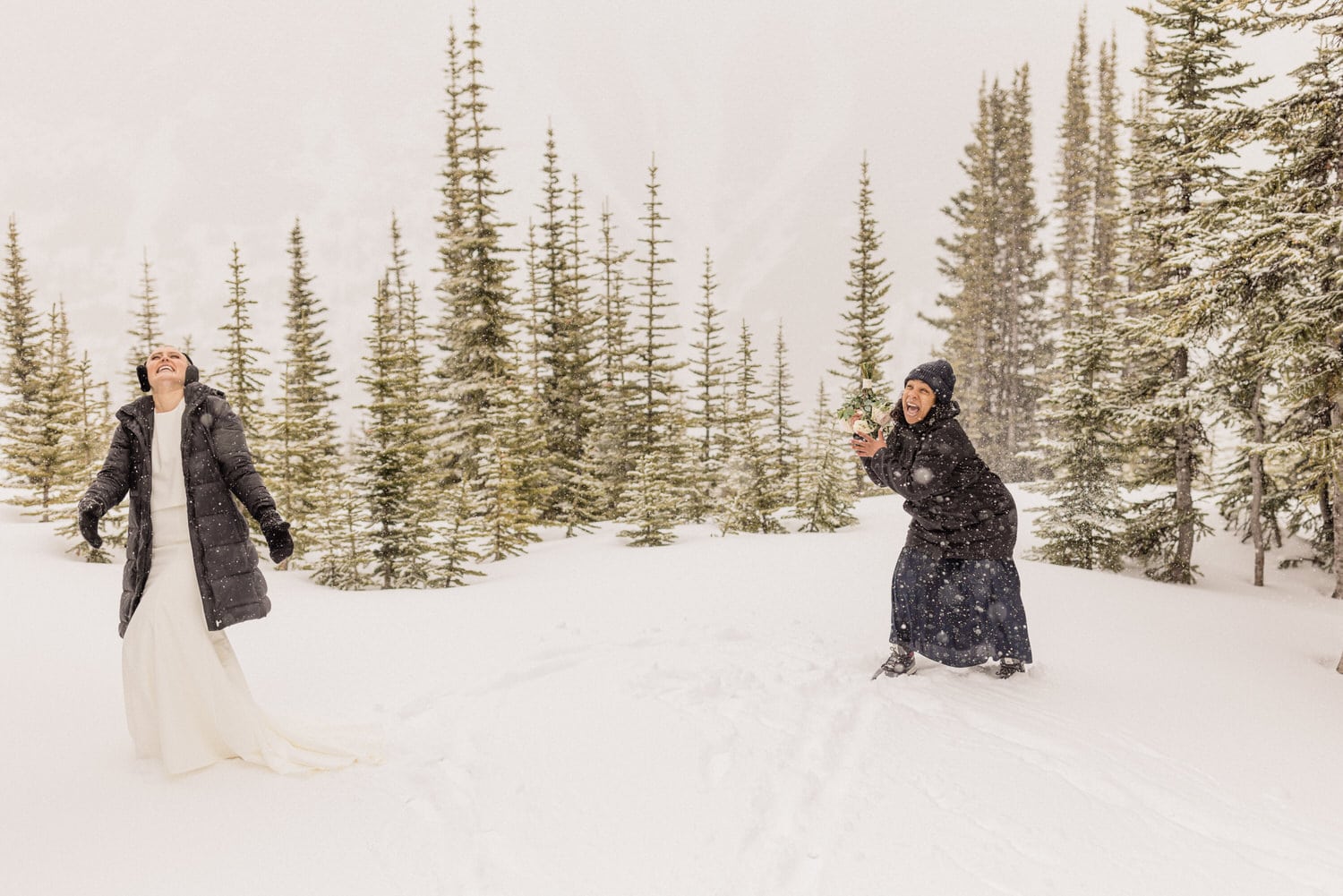 Two women enjoy a snowy landscape, one in a wedding dress and the other in winter attire, surrounded by evergreen trees while snowflakes gently fall.