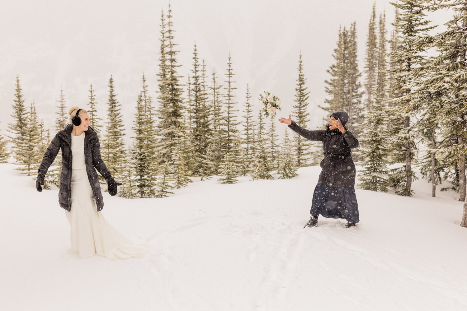 A bride in a white dress and a winter coat joyfully interacts with a friend in a black coat, amidst a snowy landscape with evergreen trees.