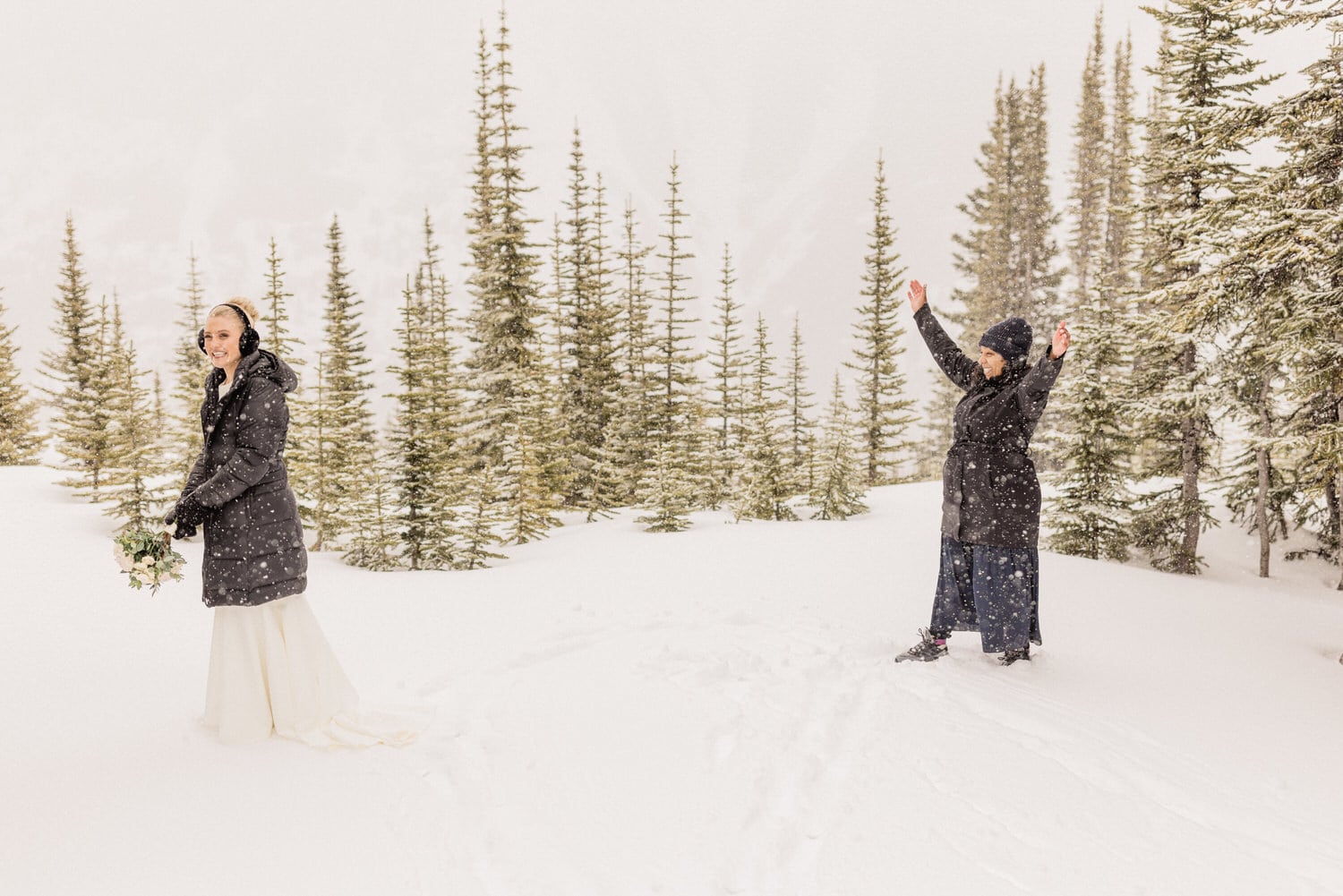 A joyful bride stands in a snowy landscape, holding a bouquet, while a guest cheers with raised hands in the background amidst evergreen trees.