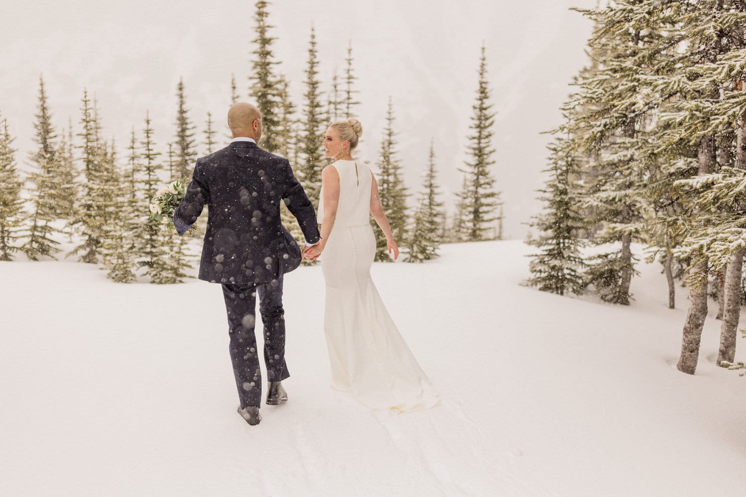 A couple walking hand in hand through a snowy landscape, adorned with evergreen trees, during their winter wedding ceremony.