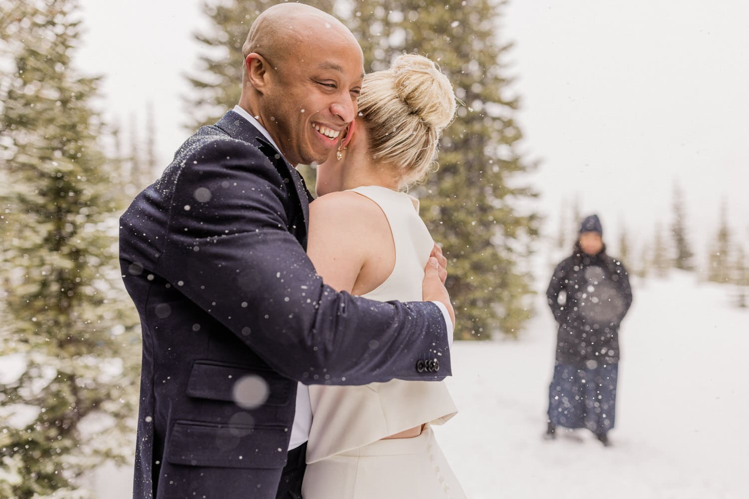 A couple embraces joyfully in a snowy landscape, celebrating their wedding amid falling snow, with a figure in the background.