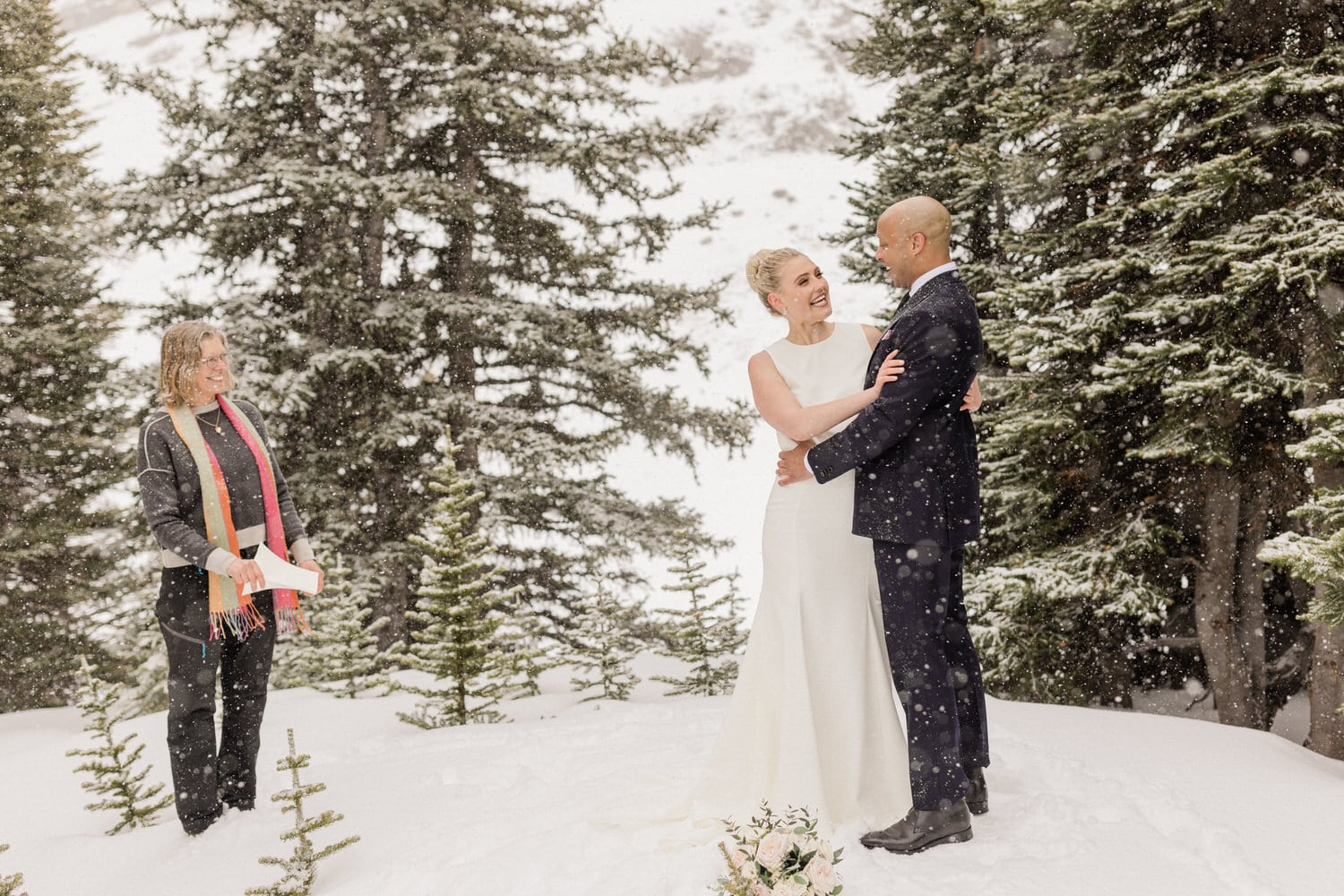 A couple exchanging vows in a snowy forest, with a celebrant standing nearby, creating a romantic and picturesque scene.