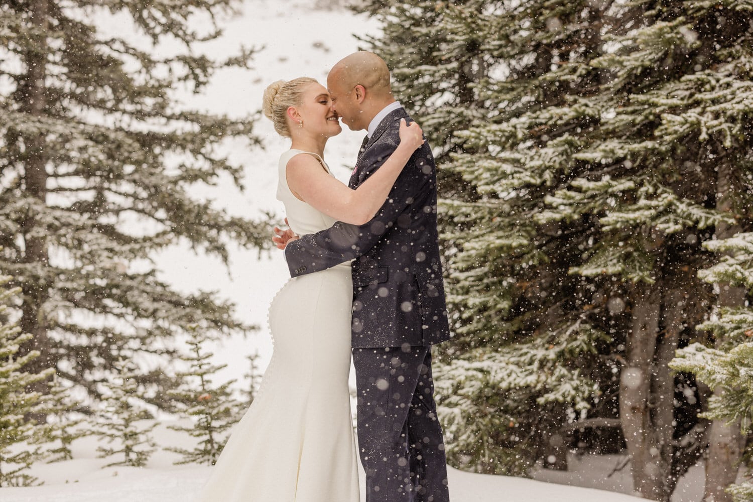 A couple sharing a loving moment while embracing in the snow, surrounded by evergreen trees.