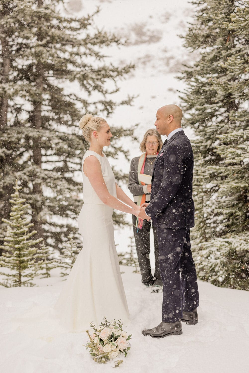 A couple stands hand in hand during their wedding ceremony in a snowy landscape, accompanied by an officiant and surrounded by evergreen trees. A bouquet is placed in the snow nearby.