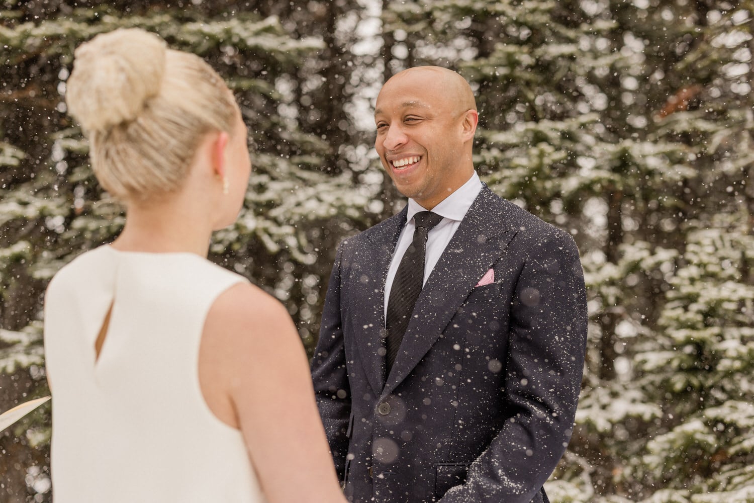 A couple exchanges joyful smiles and vows amidst falling snow in a serene forest setting.