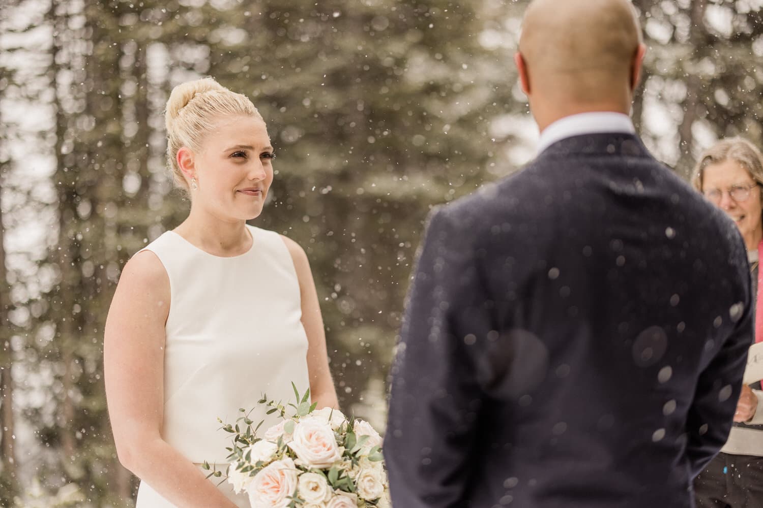 A bride with a bouquet stands smiling during her wedding ceremony in a snowy outdoor setting, while the groom faces her, and a officiant looks on.