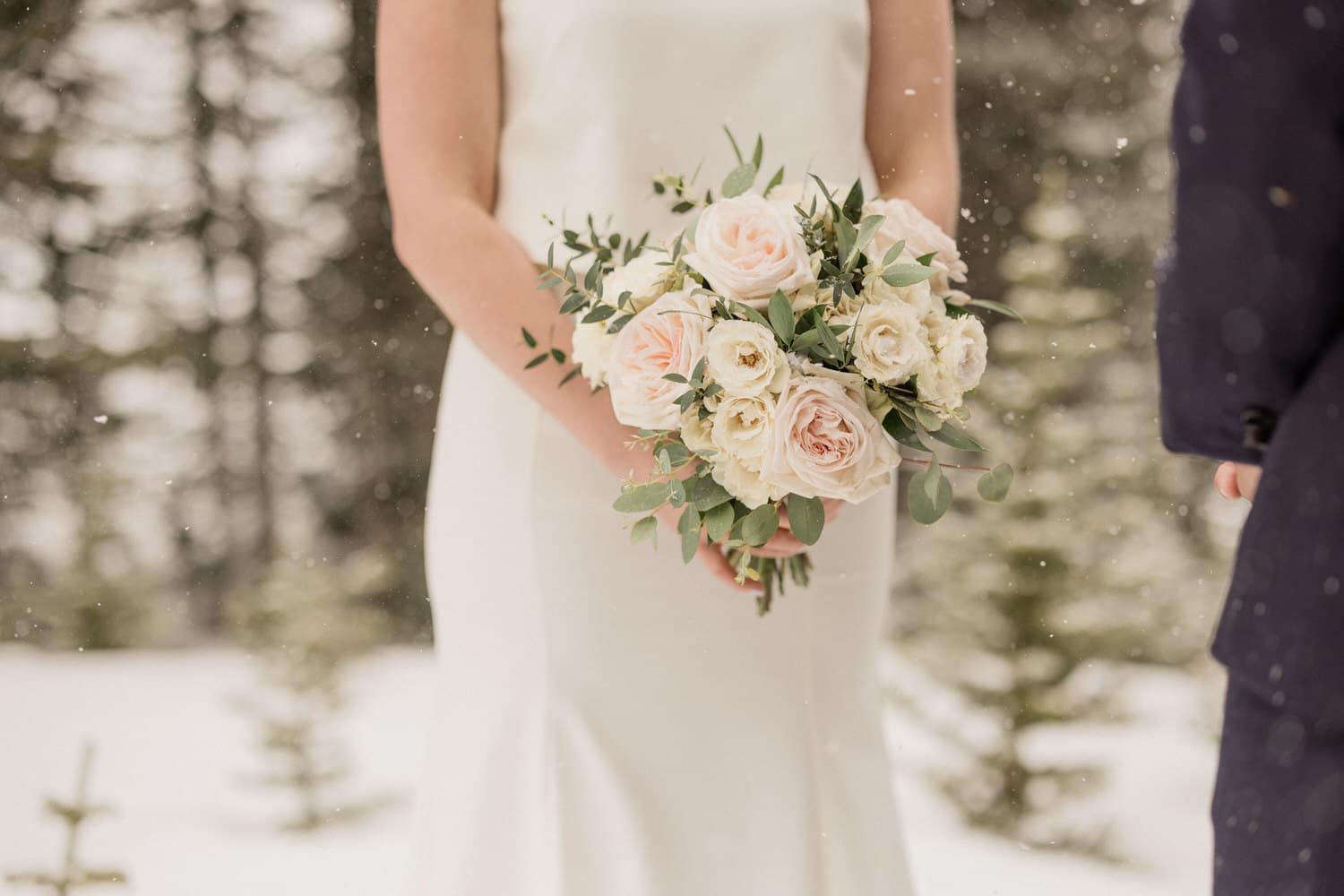 Bride holding a bouquet of pale pink and white roses in a snowy forest setting.