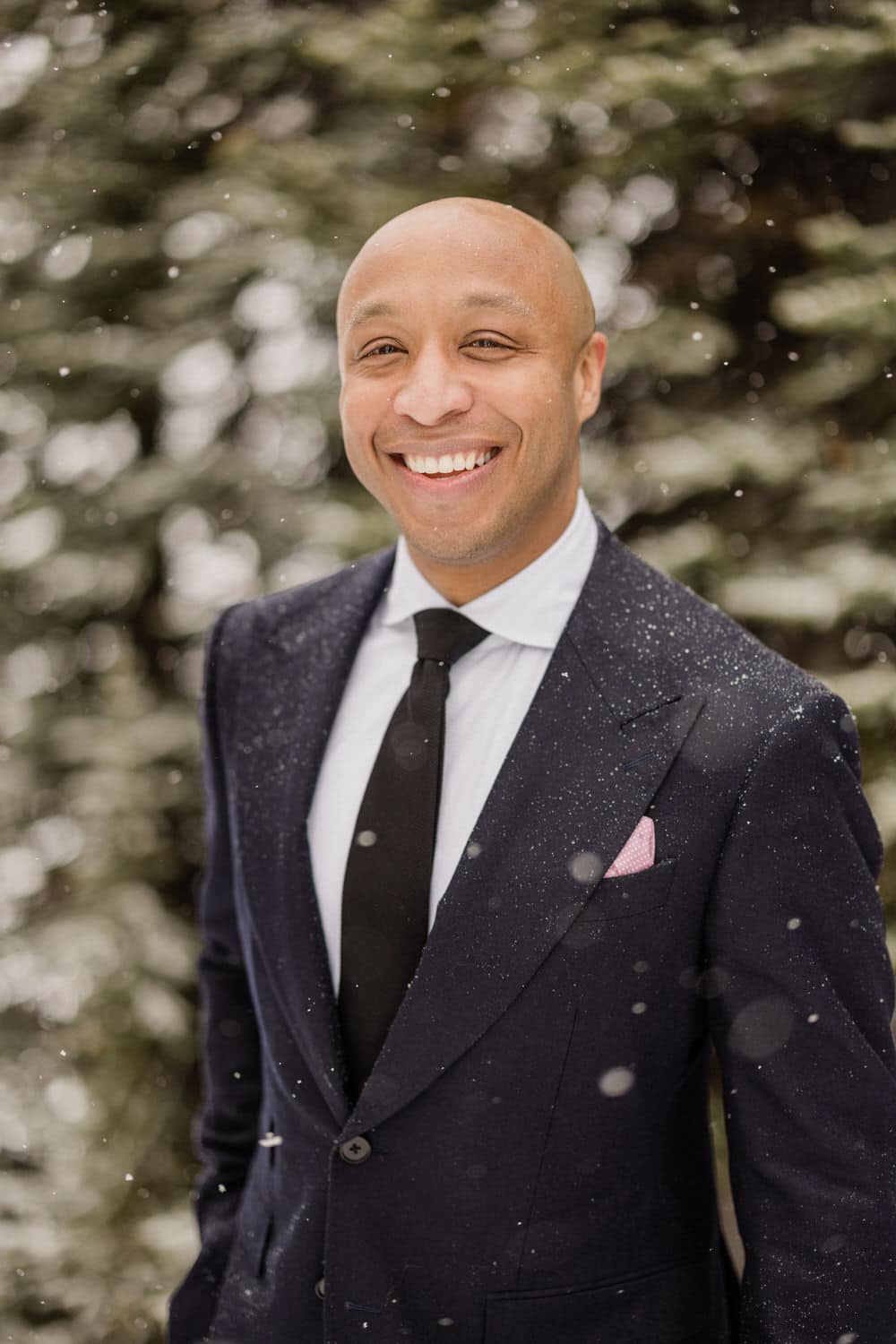 A smiling man in a formal suit standing outdoors in falling snow, surrounded by blurred evergreen trees.