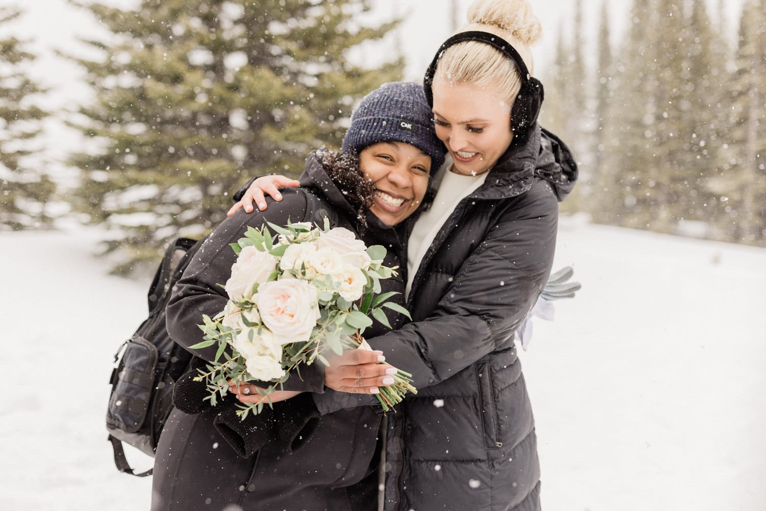 Two women embracing in a snowy landscape, one holding a bouquet of flowers, while snowflakes gently fall around them.