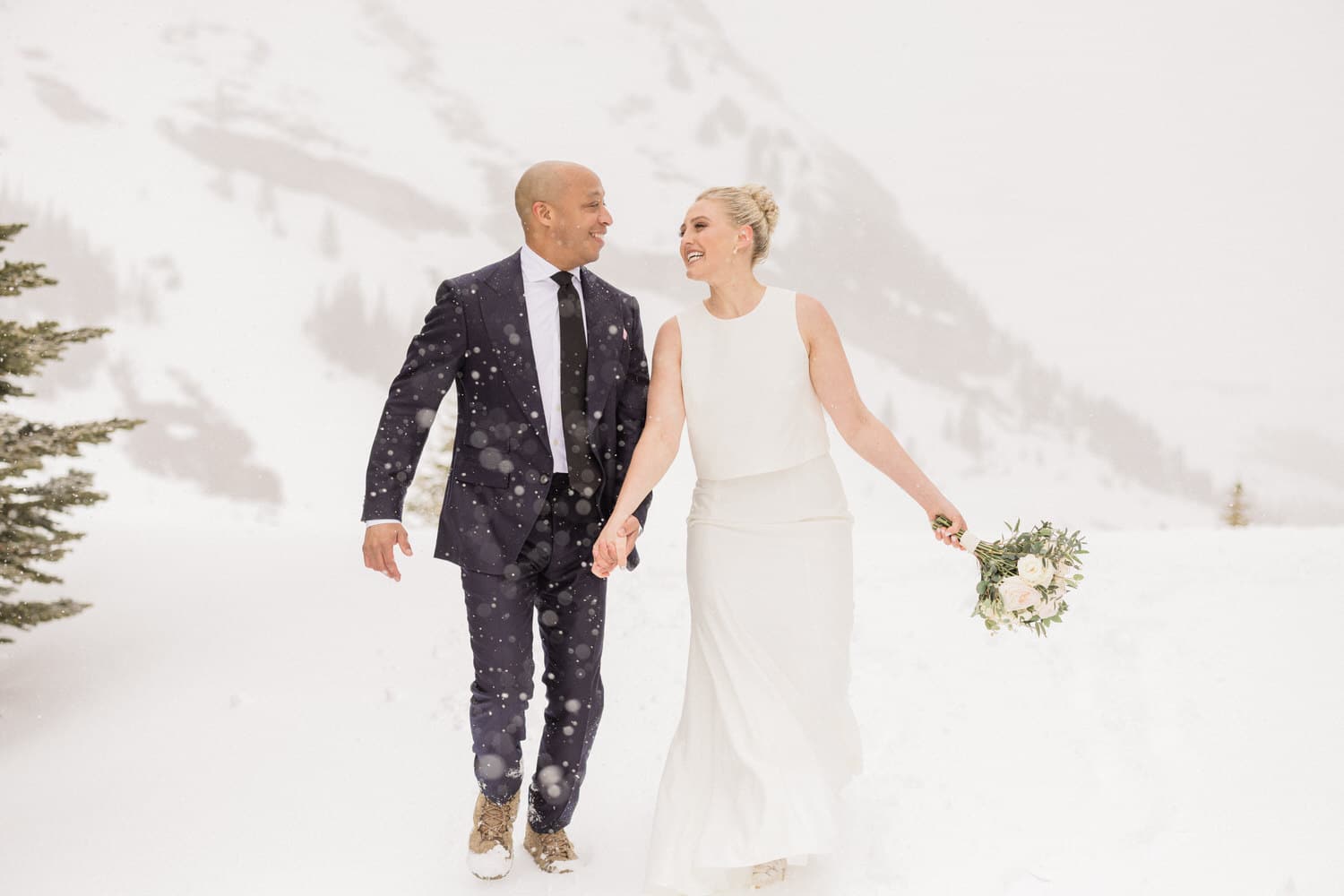 A couple joyfully walking hand in hand through the snow, surrounded by a snowy landscape, celebrating their wedding.