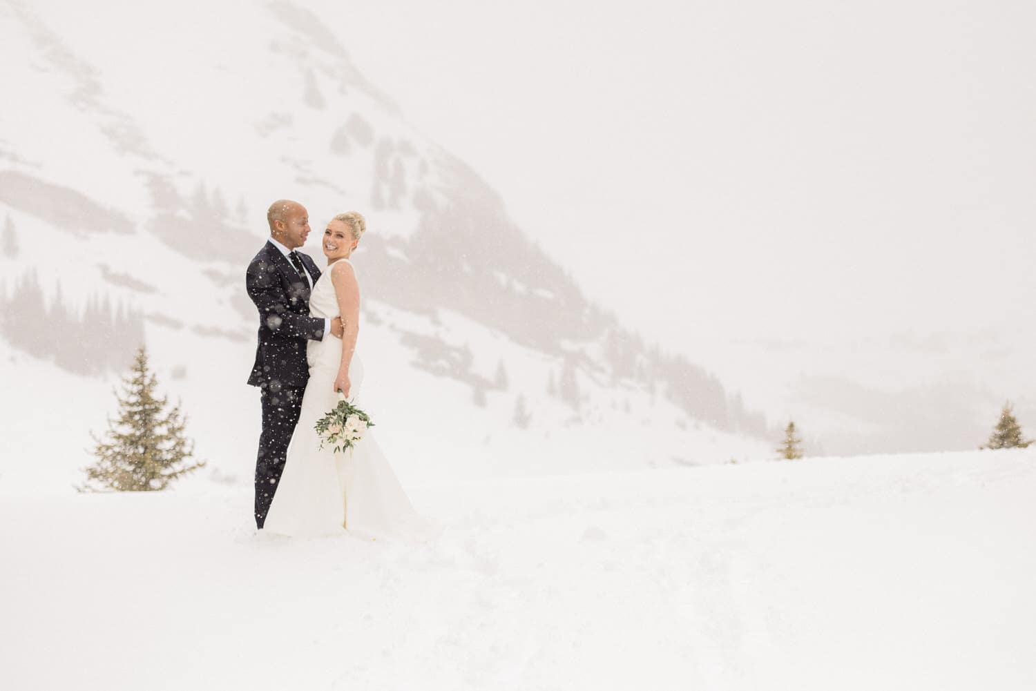 A couple stands warmly embracing in a snowy landscape, celebrating their wedding amidst falling snowflakes and majestic mountains.