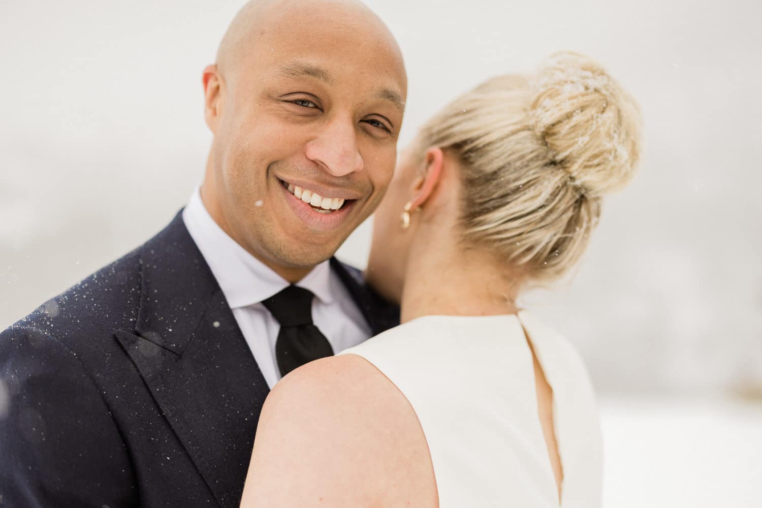 A joyful couple embracing in the snow, with the man smiling broadly and the woman leaning in closely, showcasing love and warmth amidst a winter backdrop.