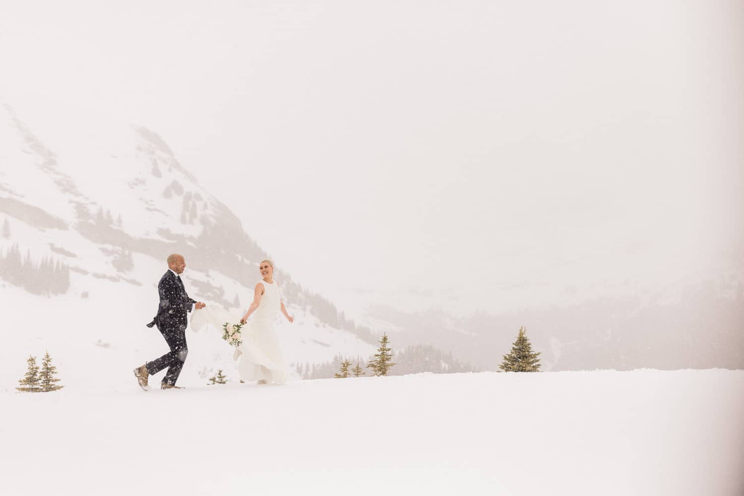 A couple joyfully walks through a snow-covered landscape during their wedding ceremony. The scene captures the enchanting backdrop of snowy mountains and trees.