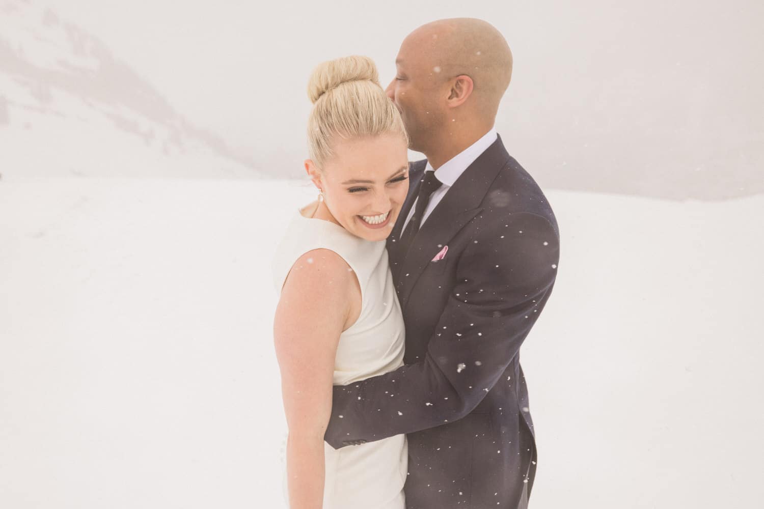 A joyful couple embracing in a snowy landscape, with the woman in a sleek white dress and the man in a dark suit, sharing a loving moment.