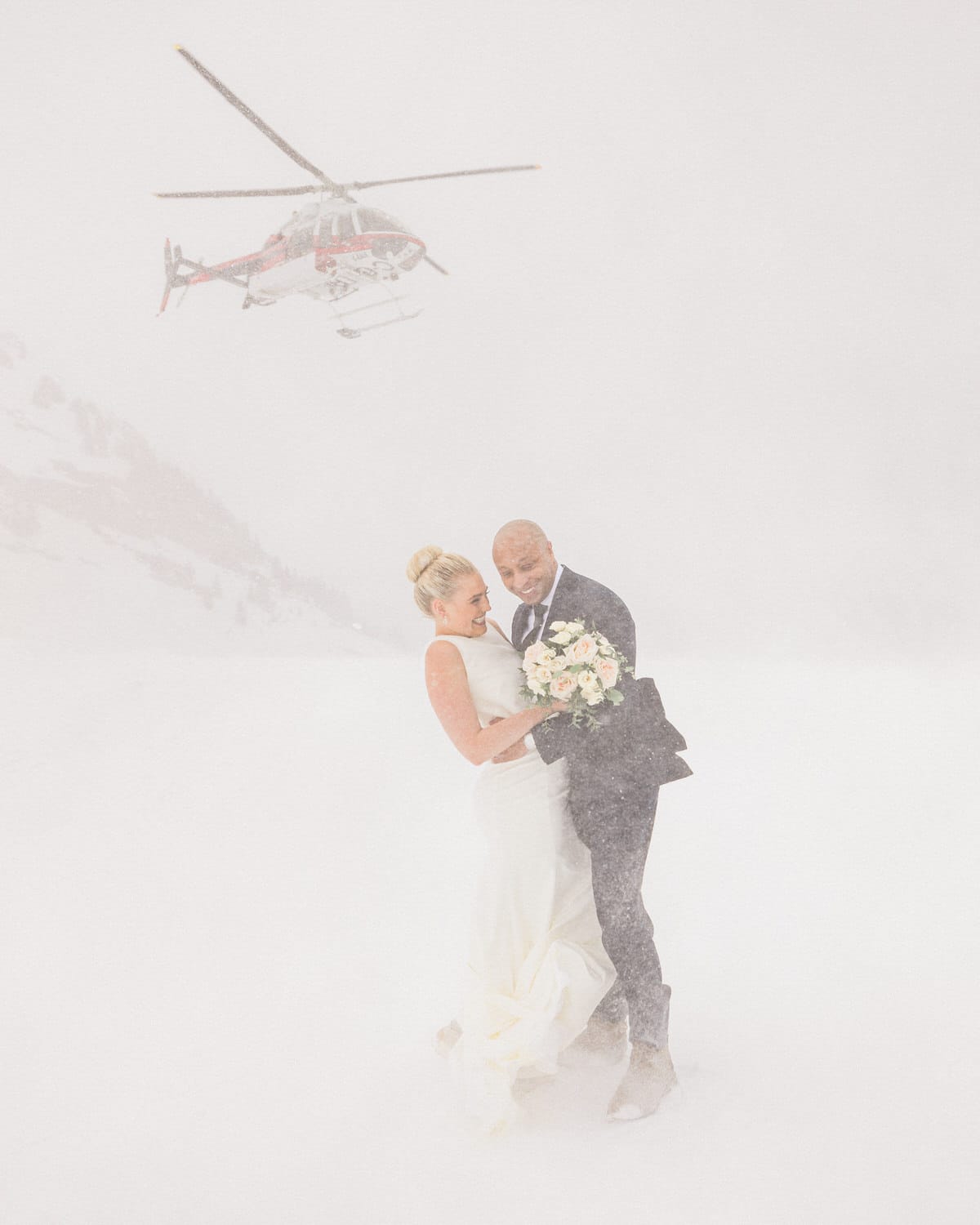 A joyful couple embraces in the snow during a wedding, with a helicopter flying above amidst a snowy backdrop.